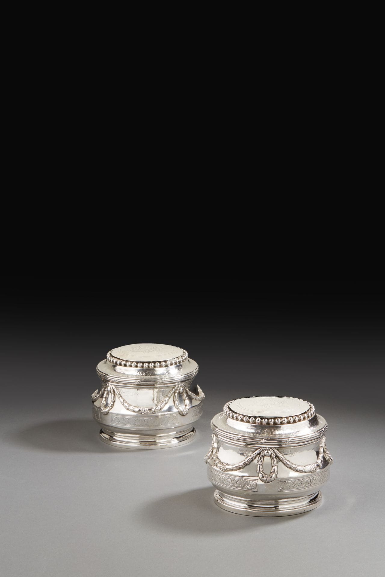 PARIS 1779 - 1780 et 1780 - 1781
Paire de grandes boîtes de toilette de forme ronde, reposant sur une bâte. La partie inférieure du corps reçoit un décor ciselé dans quatre réserves, d’un motif d’enroulements inversés de feuilles et de fleurs. Le corps est rehaussé de guirlandes de lauriers en chute maintenues par des rubans. Le couvercle mobile à doucine, mouluré de filets enrubannés, est ciselé d’un décor rappelant l’esprit du motif de la base. La bordure supérieure de la doucine est agrémentée d’un collier de perles. Gravée aux armes de la famille Church, originaire d’Earls Colne dans le Comté d’Essex, Grande-Bretagne.
Maître orfèvre : François JOUBERT, reçu en 1749
Poinçons : pour les corps lettre Q, pour les couvercles lettre S. Probablement inversion des couvercles au cours d’un partage antérieur.
Poids : 1337,5 g - diamètre : 13 cm - hauteur : 10,8 cm

Provenance :
Vente PAULME du 3 mai 1909, sous le numéro 11, et vente à Versailles du 8 décembre 1974, sous le numéro 98, en première de couverture
PARIS 1779 - 1780 and 1780 - 1781
A pair of large toilette boxes
Master silversmith: François JOUBERT