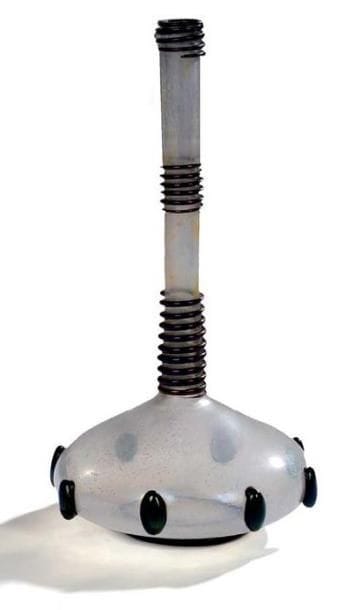 Umberto BELLOTTO (1882-1940) Designer (attribué à) & Fratelli TOSO Verrier
Vase soliflore à base renflée et aplatie. Épreuve en verre irisé nacré. Décor en application de cabochons de verre noir lustré en pourtour de la panse et de sections de serpentins, également en verre noir lustré, autour du col. Un léger manque vers l'extrémité d'un serpentin. Hauteur: 31 cm