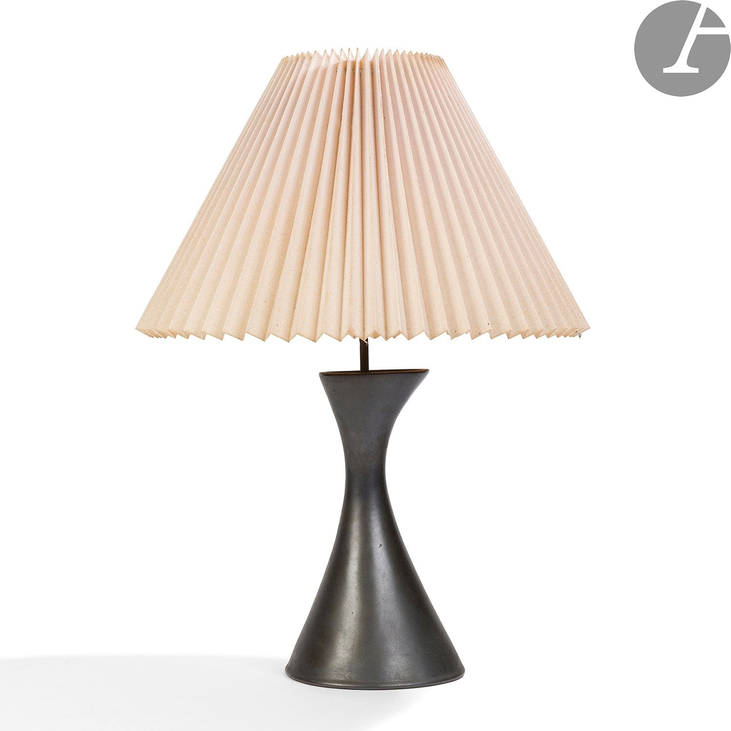 GEORGES JOUVE (1910-1964) 
Diabolo asymétrique
Rare lampe de salon.
La base titre en céramique émaillée noir ; le fût central en laiton et scellé au plâtre fin d’origine.
Système électrique et support d’abat-jour réglable en hauteur d’origine.
Abat-jour d’origine en tissu cartonné plissé ivoire.
Années 1950.
Le pourtour du trou de passage légèrement repris à l’origine, une brûlure et manque sur le support d’abat-jour, légères déformations et salissures à l’abat-jour.
Signé JOUVE et du monogramme A. P. (aux lettres entrelacées) pour Apollon, marque dite à l’Alpha, au revers. 
Hauteur céramique seule : 27,5 cm 
Hauteur totale : 56 cm

Provenance :
Collection particulière, Vosges.
