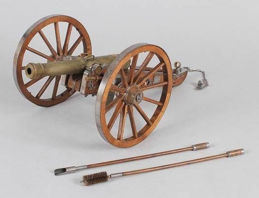 Maquette de canon système Vallée. Fût en métal patiné à deux anses et deux tourillons avec plaque en laiton frappée "Meudon 1884". Roues à rayons en bois, cerclées de fer. Long.: 70 cm. - Haut.: 30 cm. Avec ses accessoires, sceau, écouvillon et refouloir. B.E. Reproduction espagnole du XXe siècle.