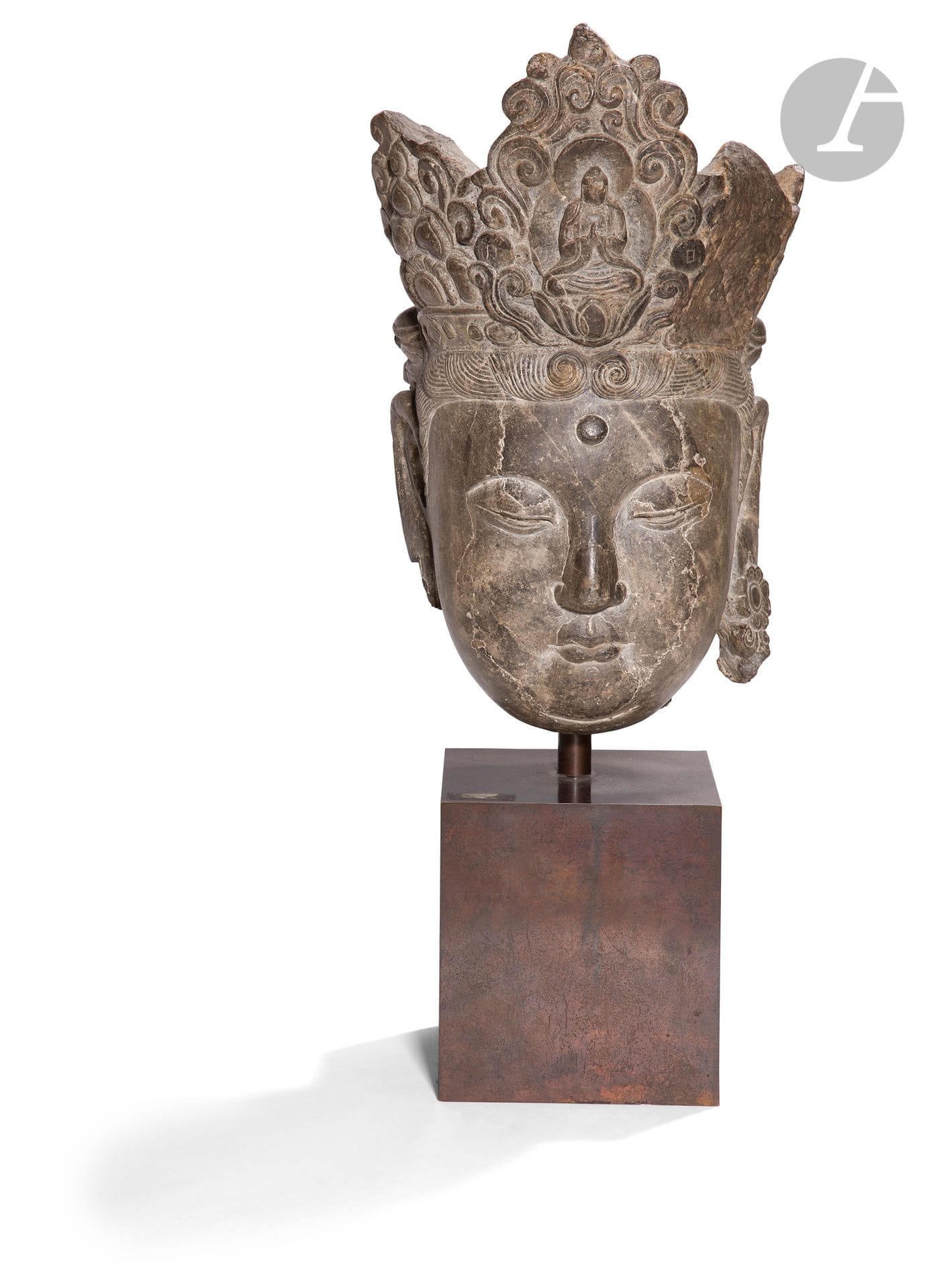 * Tête de Guanyin en pierre calcaire, Chine, époque Ming, XVe siècle
Le visage ovale sculpté avec une expression sereine et méditative. Les sourcils hauts et en arc de cercle dont l’arête est très marquée, sur des yeux plissés presque fermés. Les yeux surmontés de paupières lourdes et très renflées. Un urna (symbole de la vision dans le monde divin) en relief orne le milieu du front. Les lèvres bien sculptées et charnues se terminent en pointe au niveau de la bouche renforçant le côté méditatif du visage. Le menton est accentué par une incision en arc de cercle. Les lobes d’oreilles sont allongés décorés de petites fleurs faisant office de boucles d’oreilles. Le front est parcouru de mèches arrondies encadrant la totalité du visage jusqu’au bas des oreilles tandis que le reste est peigné en chignon sur le dessus de sa tête. La tête est scindée d’une haute tiare, sculptée en relief en son milieu d’une figure du Bouddha assis en Namaskara Mudra sur un lotus et entouré d’une mandorle. Autour des volutes en haut relief et des pétales de lotus stylisés ornent le reste de la coiffe. De chaque côté du bas de la mandorle au milieu des volutes un médaillon rond est gravé d’un idéogramme, à gauche ÈÕ (ri) - le soleil, à droite 月(yuè) - la lune.
Hauteur : 35 cm
Accidents sur le côté gauche de la coiffe, au bas de l’oreille droite et à l’arrière de la tête. Peut-être quelques petits rebouchages.