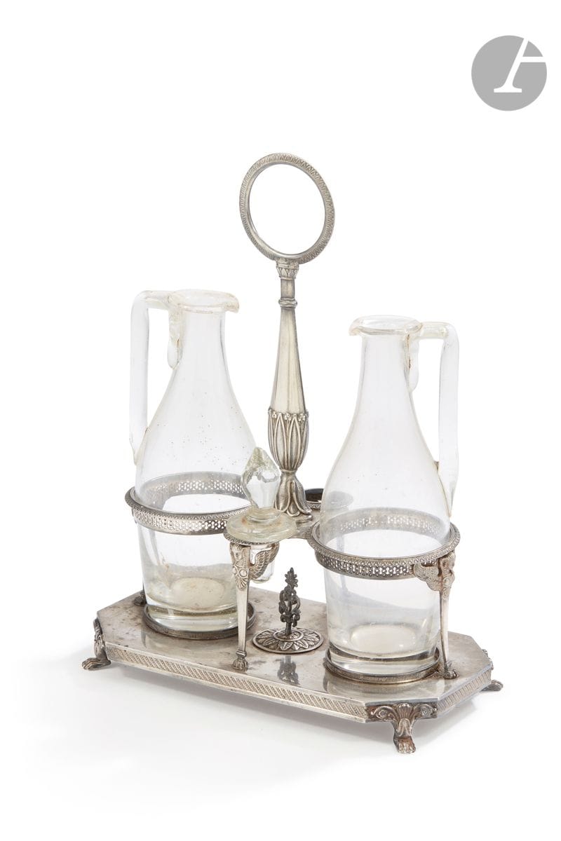 PARIS 1809 - 1819
Huilier-vinaigrier en argent et ses carafes reposant sur une terrasse rectangulaire à pans coupés posée sur quatre pieds griffe, les supports des carafes en sphinges ailées. La prise centrale est rehaussée de fleurs de lotus et de feuilles d’eau.
Orfèvre : Sixte-Simon RION, insculpe en 1807
Poids : 677 g - hauteur : 30 cm
État : manque un bouchon de verre