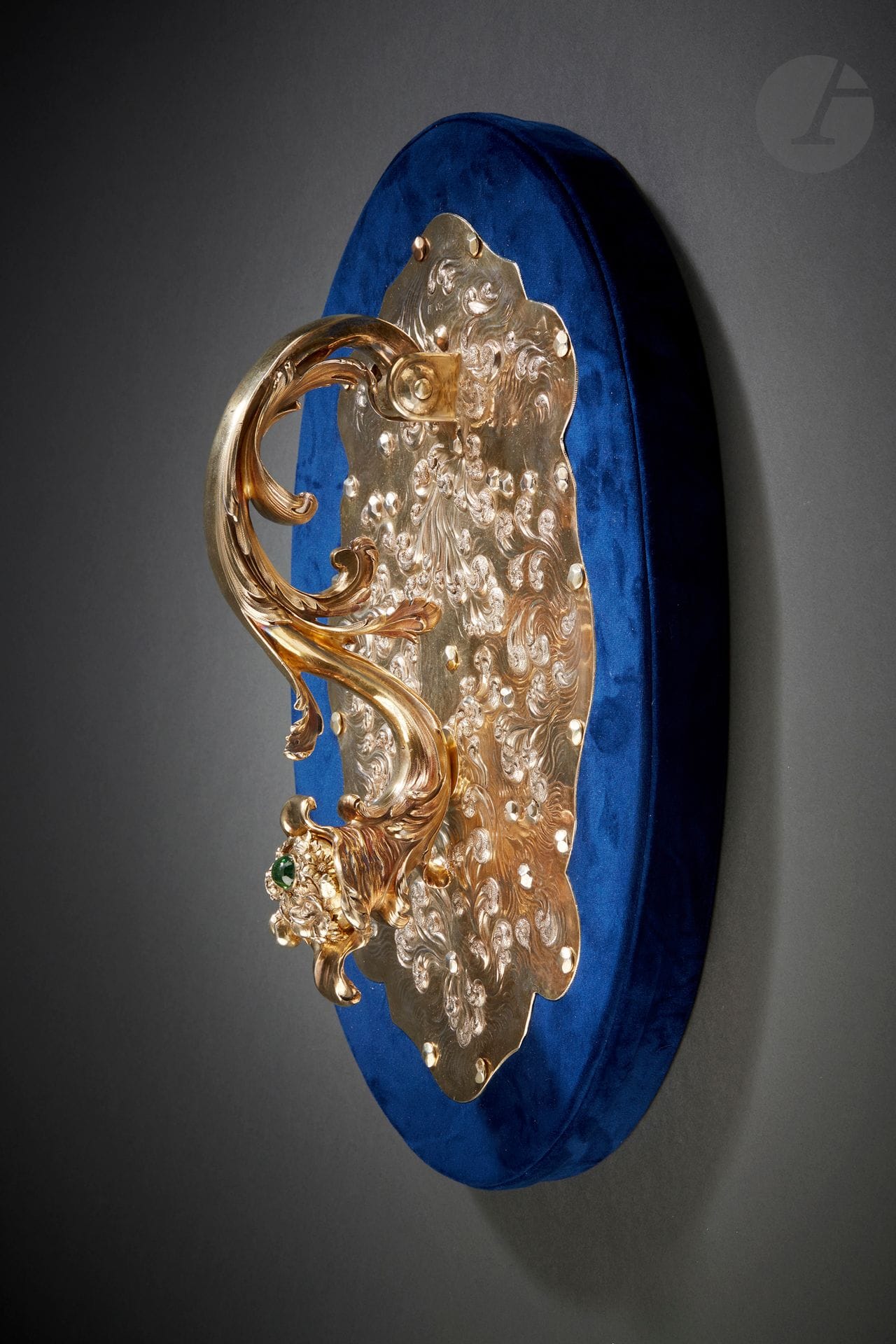LONDRES VERS 1830
Heurtoir en vermeil rehaussé d’un cabochon de tourmaline verte reposant sur un socle ovale recouvert de velours bleu. Le support de forme ovale à contours est décoré de vagues sur fond amati dans un mouvement tourbillonnant. Le heurtoir en forme de S agrémenté de feuilles d’acanthe est terminé par une corne fleurie.
Orfèvre : MORRIS & MICHAEL EMANUEL, insculpe en 1825
Poids brut : 5536 g - hauteur du heurtoir : 38 cm - largeur : 24 cm