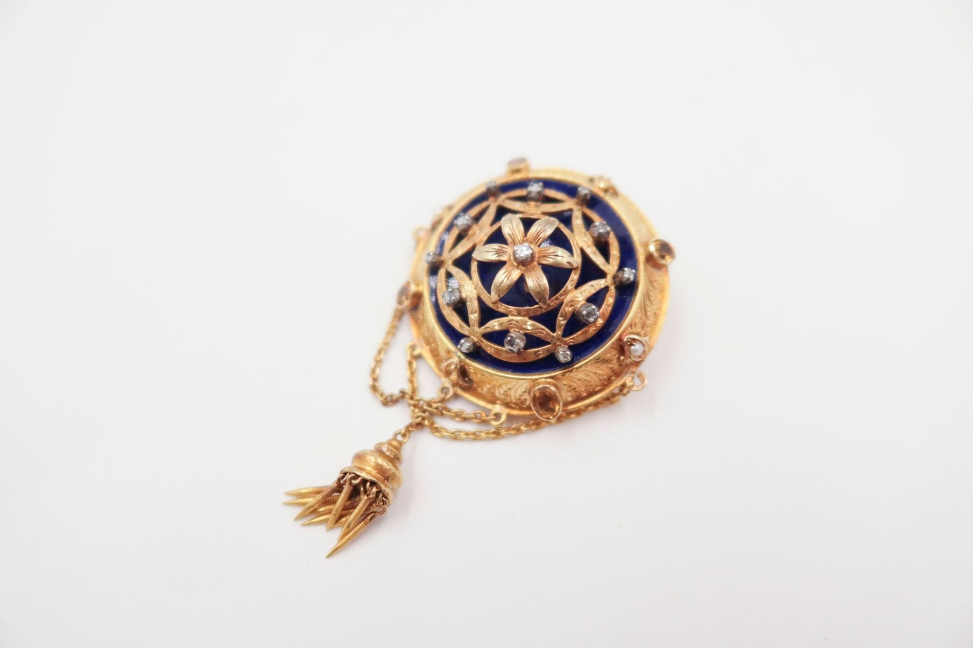 Round brooch in 18K (750) gold, the blue enamelled background decorated with gold flowers set with rose-cut diamonds, the setting set with yellow stones and half pearls. Work of the last quarter of the 19th century. Gross weight: 17 g (missing)