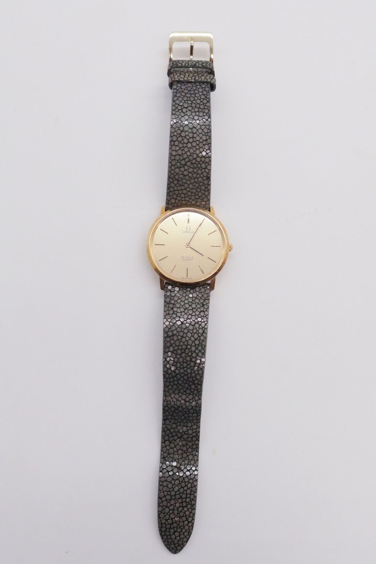 OMEGA DE VILLE
Montre bracelet en or jaune 18K (750) sur cuir. Boîtier rond. Cadran doré avec index bâton appliqués. Mouvement quartz. Bracelet galuchat avec boucle ardillon rapportée. 
Cadran et boîtier signés. 
Numérotée 1910044.
Diam. : 34 mm
Poids brut : 40 g
(Microrayures d'usage)