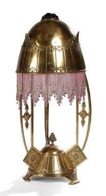 TRAVAIL ALLEMAND
Importante lampe en laiton, en partie doré, à décor repoussé dans le goût néoclassique. L'abat-jour en obus, à la terminaison d'un cabochon façon verre taillé, présente un rideau perlé en pourtour. Hauteur: 53 cm