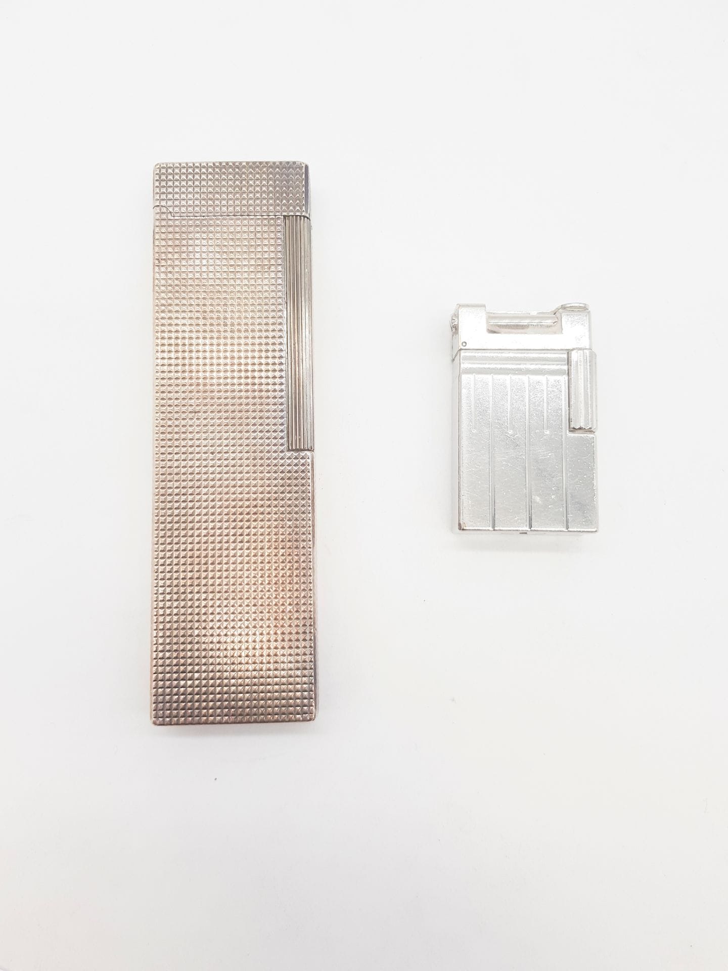 DUPONT 
Lot de deux briquets en métal argenté, l'un, de table, guilloché, l'autre à décor de stries verticales et inscription gravée "PERSPECTIVE 2000". Hauteur briquet de table : 14 cm environ (usures, fonctionnement non garanti)