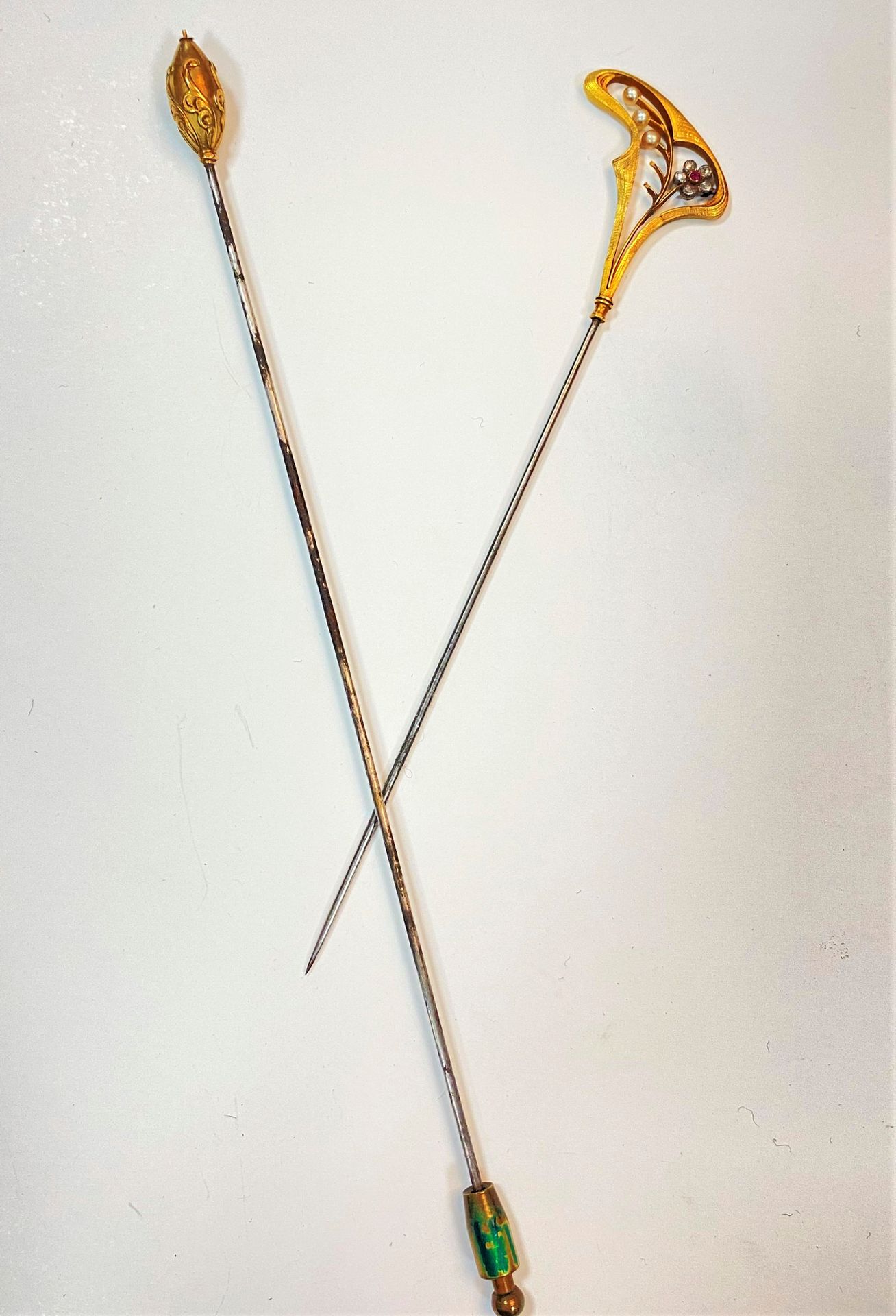 Lot de deux épingles en métal et or 18K (750) l'une sommée d'un motif ajouré et guilloché agrémenté d'une fleur sertie d'un rubis, les pétales en diamants taillés en roses, le feuillage agrémenté de perles, l'autre sommée d'un motif ovale à décor de feuilles d'acanthe. Travail français du début du XXe siècle. Poids brut : 11,2 g (manques)