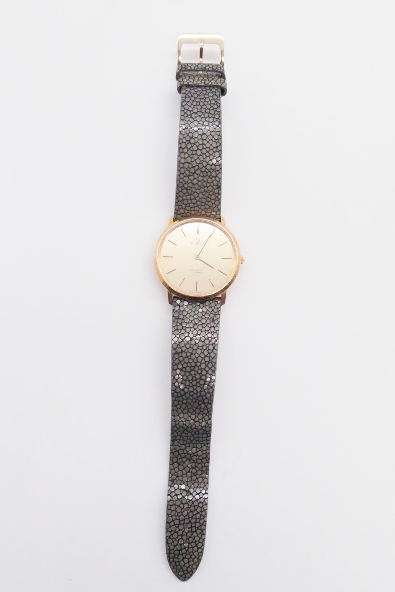 OMEGA DE VILLE
Montre bracelet en or jaune 18K (750) sur cuir. Boîtier rond. Cadran doré avec index bâton appliqués. Mouvement quartz non signé et rapporté. Bracelet galuchat avec boucle ardillon rapportée. 
Cadran et boîtier signés. Numérotée 1910044.
Diamètre : 34 mm
Poids brut : 40 g
(Microrayures d'usage)