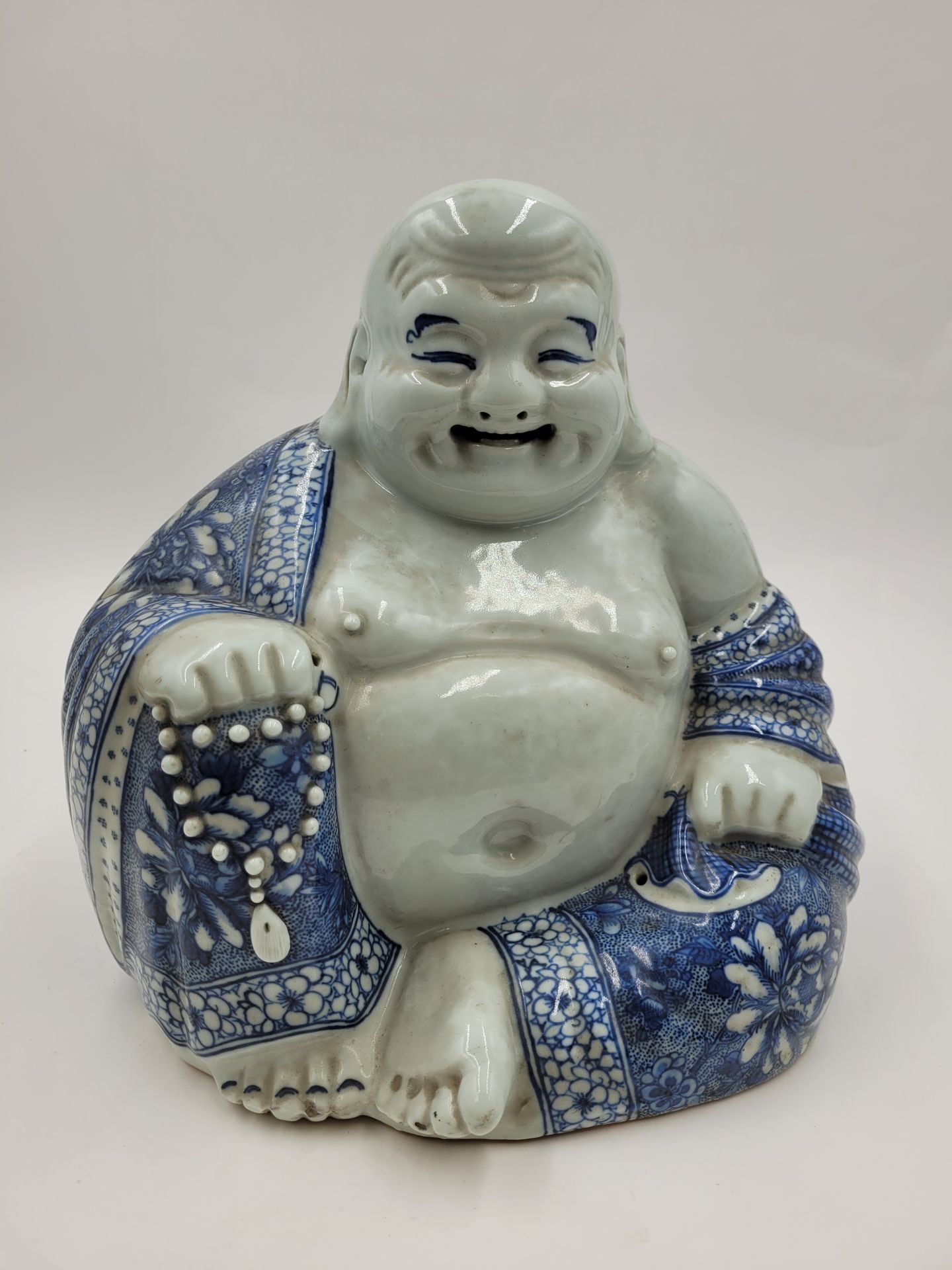 Putaï Bouddha rieur, Chine, début XXe 
En porcelaine à décor bleu blanc, manteau fleuri et chapelet dans la main droite. 
Hauteur : 26,5 cm 
Petits éclats aux orteils.