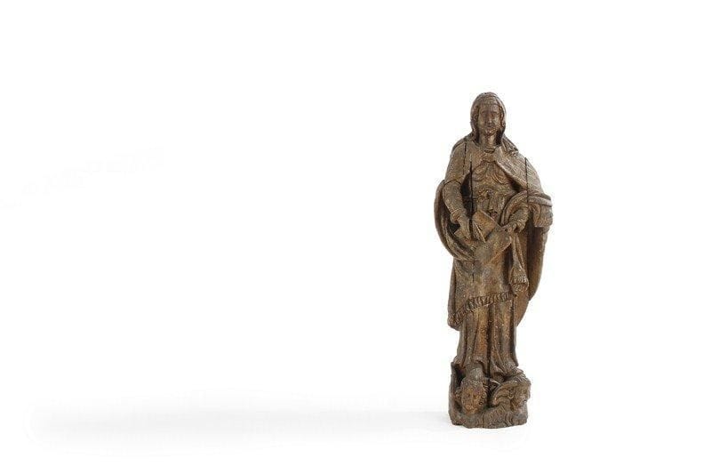 Sainte femme en bois sculpté du début du XVIIIe siècle. Elle présente le Livre saint de sa main gauche et désigne une page de la droite, elle est figurée debout avec des chausses en forme de patte d'ours qui reposent sur deux têtes d'anges ailés. Importantes fentes verticales sur toute la hauteur et nombreux petits manques. H : 85 cm.