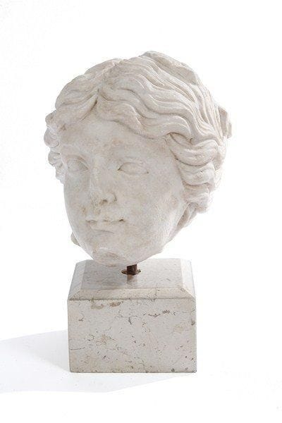 Dans le goût de l'antique. Tête de femme en marbre blanc de Carrare sur un socle en pierre. Manques. H : 21 cm.