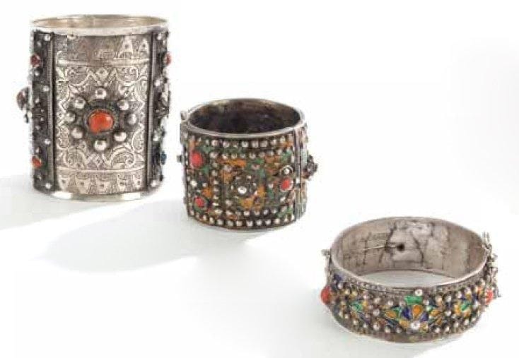 CHEVILLIÈRE ET DEUX BRACELETS EN ARGENT, ALGÉRIE, BENNI YENNI, XXE SIÈCLE Chevillière à charnière gravée d'un décor fleuronné et larges cabochons de corail et bracelets ciselés agrémentés de perles en argent et cabochons de corail, sur fond émaillé en jaune, vert et bleu. Chevillière: H.: 10 cm et diam.: 8,5 cm; et bracelets: diam.: 6 et 5,3 cm