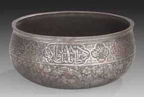 BASSIN TAS EN BRONZE, IRAN SAFAVIDE, XVIIE - XVIIIE SIÈCLE Bassin à fond plat et panse bombée. Décor gravé sur la paroi extérieure d'un bandeau épigraphié thuluth sur fond hachuré donnant une invocation religieuse en partie haute. Sur la moitié inférieure, rinceaux de demipalmettes formant des entrelacs géométriques, sur fond hachuré également. Plusieurs marques de possesseurs sous le fond, dont une dans un médaillon en mandorle donnant «Le propriétaire est Husayn Aghâ Bâbâ». Diam.: 22,8 cm
