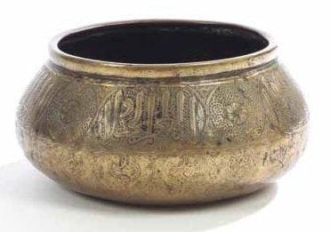 BASSIN TAS EN LAITON, IRAN, ART QÂJÂR, XIXE SIÈCLE Bassin à base bombée, paroi convexe et ouverture cintrée, à décor gravé et traces d'incrustation en pâte noire. Paroi extérieure ceinturée d'un large bandeau composé de cinq cartouches épigraphiques thuluth à la dédicace anonyme, alternés de médaillons circulaires à rosette et rinceau spiralé. Sous ce bandeau, frise de larges lambrequins et palmettes lancéolées alternant. Sous la base, marque du propriétaire donnant «Sâhib Mohammad Husayn». (Deux accidents) Diam.: 18 cm et H.: 11,5 cm