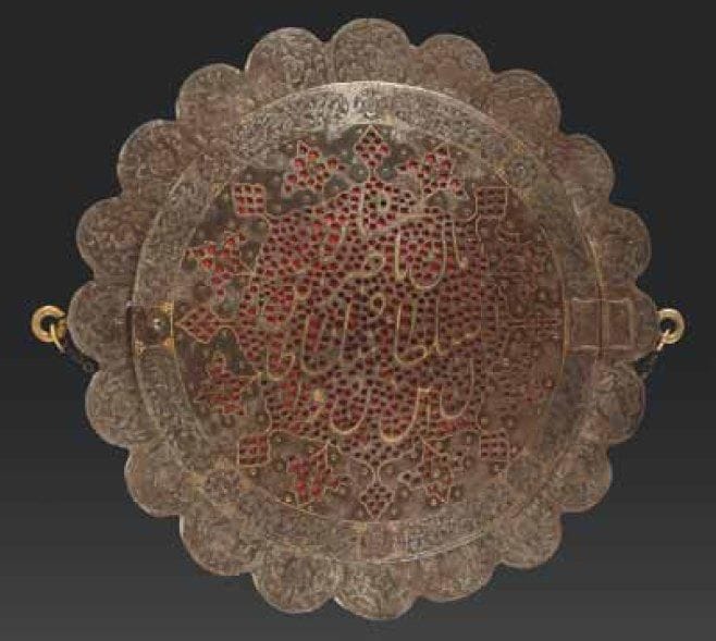 MIROIR QÂJÂR EN ACIER AU NOM DE NÂSIR AL-DÎN SHÂH (R. 1848 - 1896), IRAN, XIXE SIÈCLE Boîtier en disque polylobé à couvercle circulaire fermant par un crochet et avec deux anneaux latéraux reliés par une chaînette. Sur le couvercle, décor incrusté d'or donnant le nom du Shâh en nasta'liq dans un médaillon à pendentifs à fond de résille. Au revers, mamelon en forme de soleil rayonnant sur fond de personnages assis. Diam.: 26 cm