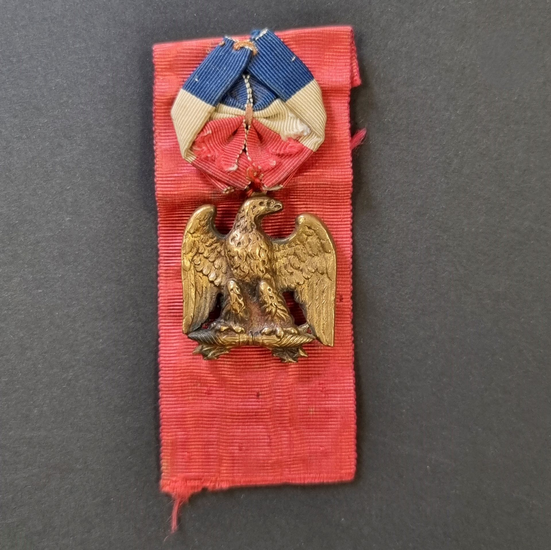 FRANCE 
Imperial Eagle veteran badge, on lightning. 
Brass, sewn to a scarlet ribbon with tricolor rosette, with button attachment. 
30 x 30 mm 
T.T.B.