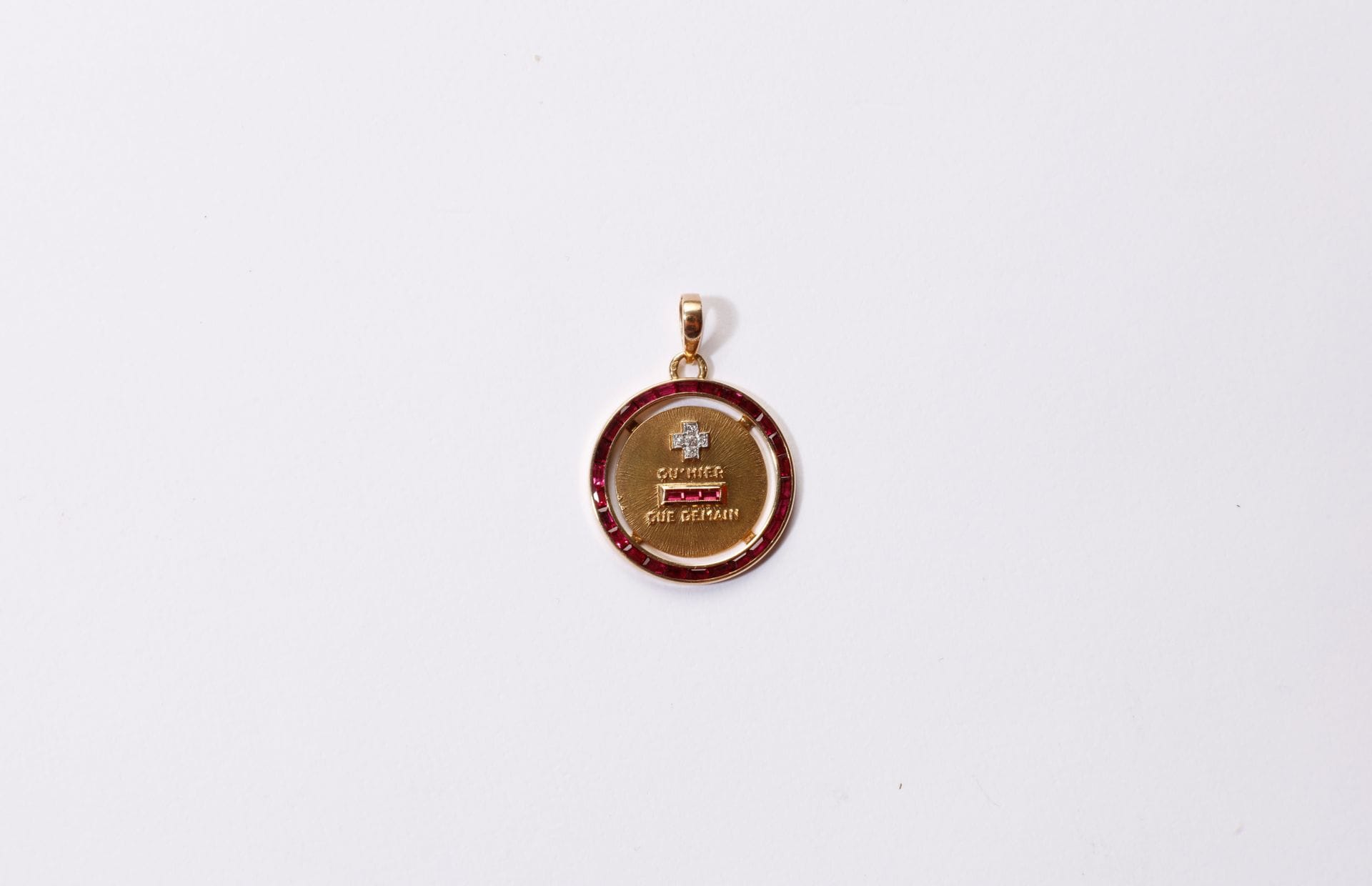 Médaille en or 18K (750) texturé portant la mention "+ qu'hier - que demain", piquée de diamants ronds 8/8, sertie de rubis calibrés. Diamètre : 2,3 cm environ. Poids brut : 3,8 g