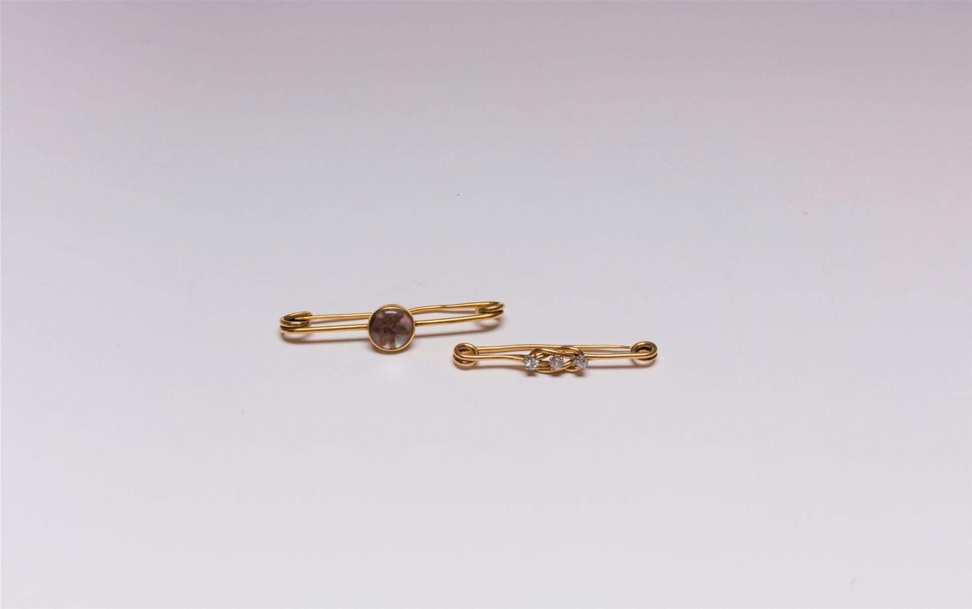 Suite de deux épingles de nourrice en or 18K (750), l'une sertie de 3 diamants ronds de taille ancienne, l'autre, ornée d'une fleur séchée sous verre galbé et sur fond de nacre (porte un numéro). 
Poids brut : 14,3 g