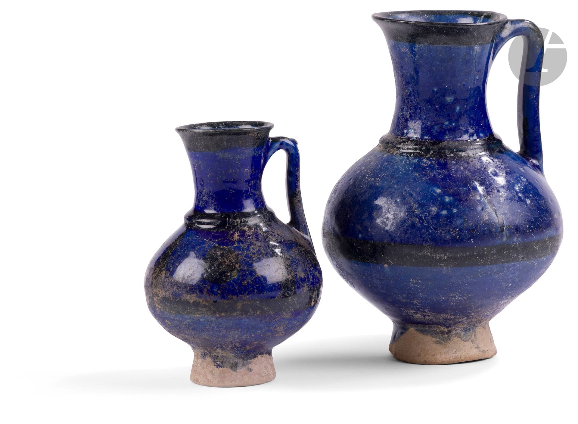 Deux pichets en céramique à glaçure bleu nuit, Iran, XIIe - XIIIe siècle
Une grande et une petite verseuse sur haut piédouche à panse globulaire surmontée d’un col cylindrique à lèvre éversée et relié à la panse par une anse coudée marquée d’un petit poucier sur la plus petite. En céramique siliceuse à décor peint en noir sous glaçure bleu cobalt, de bandes horizontales sur chacune, la panse de la petite verseuse ornée en plus d’une alternance de disques et de lignes verticales.
Hauteur : 24,5 cm et 17,5 cm
Restaurations, irisation, petits manques au pied de la grande.

Two Stonepaste Pitchers, Iran, 12th - 13th Century