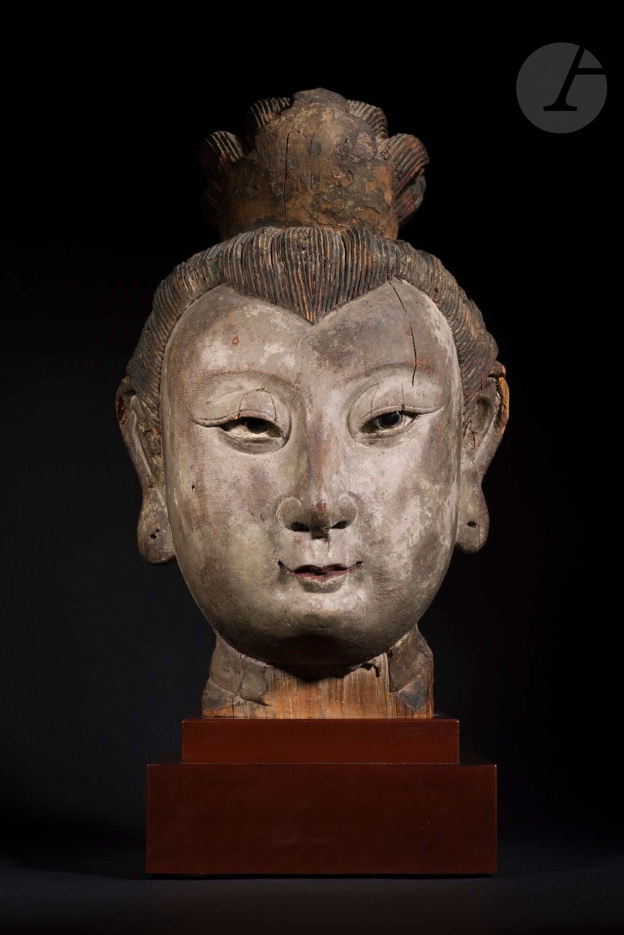 Importante tête en bois de jeune femme noble, Chine, époque Yuan - Ming, XIVe - XVe siècle
Elle porte une coiffure très élaborée tenue à l’avant et à l’arrière par un accessoire serrant le chignon. Les yeux finement sculptés sont incrustés d’une pierre noire figurant l’iris. La bouche est étroite et les lèvres fines sont relevées par une polychromie rouge. Les oreilles ont de larges lobes percés qui ont pu à l’origine être ornés de boucles. Le bois très finement sculpté est recouvert d’une gaze sur laquelle a été appliqué du gesso, très probablement pour solidifier la sculpture réalisée en plusieurs parties.
Hauteur : 46 cm ; h. avec socle : 57 cm
Manques, traces de polychromie.

Provenance : 
Acquise chez Oscar Meyer à Los Angeles entre 1950 et 1971 (copie d’une photographie).