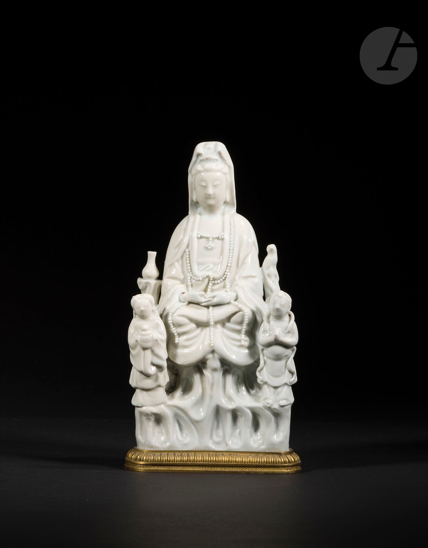 Guanyin in Chinese white porcelain, China, Dehua, 17th century
Depicted seated in dhyanasana on a rock, she is dressed in a pleated robe, open on the chest, and adorned with a long pearl necklace. Her face is serene, her eyes downcast, and her hair is gathered into a bun, topped with a tiara and covered with a cloth. She is surrounded by two stone columns on which rests a vase and a parrot. Standing at her feet on the lower part of the rock are her two acolytes LongNü, holding the Pearl of Light, and ShanCai TonZi, hands in adoration.
Height: 23.2 cm
Mounted on a gilded copper alloy base. Missing fingers.