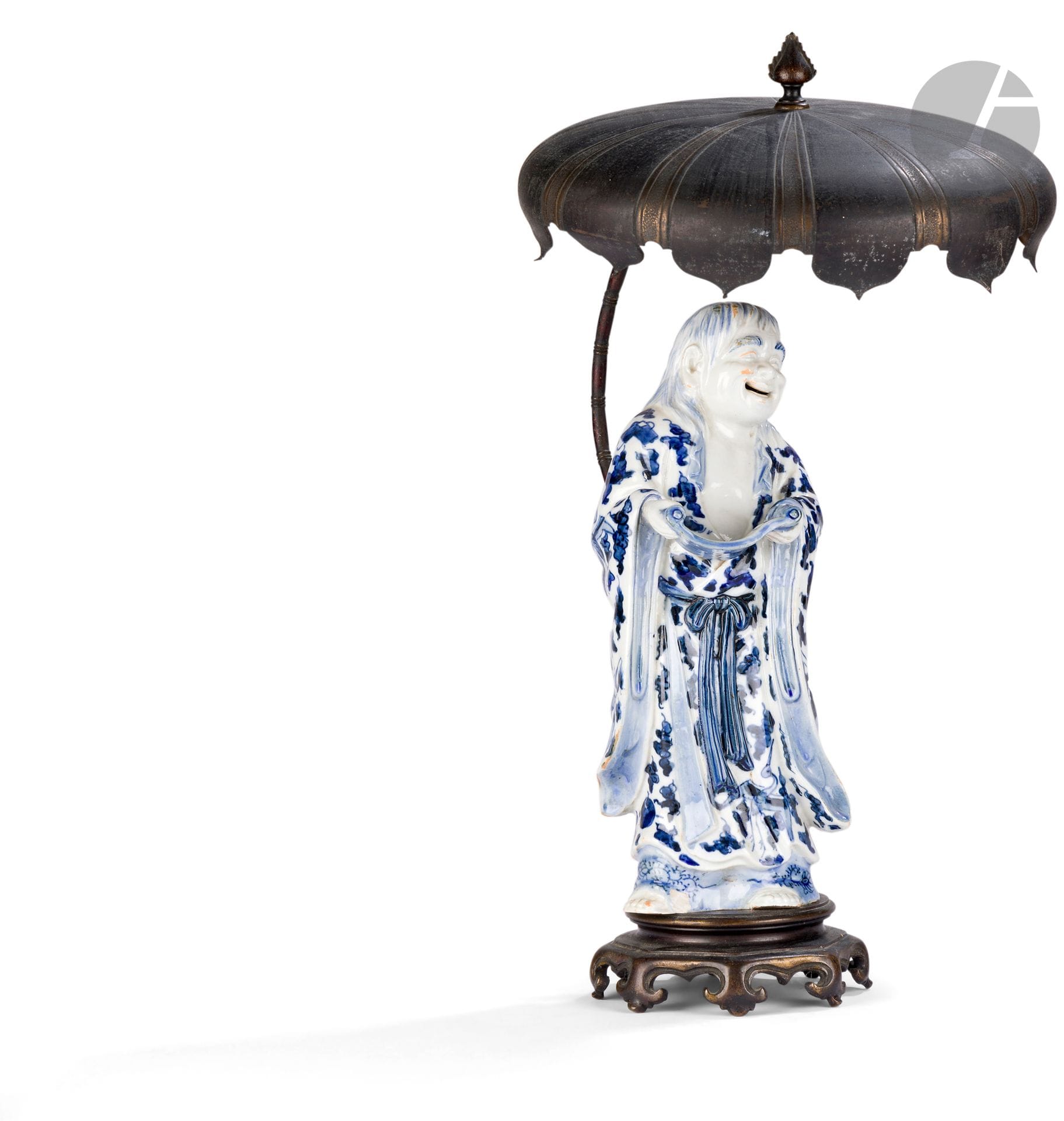 Personnage représentant Kanzan, en porcelaine bleu et blanc, monté en lampe, Japon, XIXe siècle
Kanzan, vêtu d’un kimono à décor émaillé en bleu sous couverte à décor de tsuru (grues à tête rouge), nuages stylisés et objets précieux, tient dans ses mains un rouleau calligraphié qu’il regarde avec un sourire. Il est monté sur une base octogonale à huit pieds en alliage de cuivre, à l’arrière de laquelle monte une tige courbe imitant un bambou finissant en large parasol.
Hauteur : 28,2 cm
Petit éclat.

Kanzan, dont le nom signifie « montagne froide », est un poète zen qui cherche son inspiration dans la nature. Bien qu’ils se ressemblent énormément, on le distingue facilement de son alter ego Jittoku, car il tient toujours dans ses mains un rouleau de parchemin.
