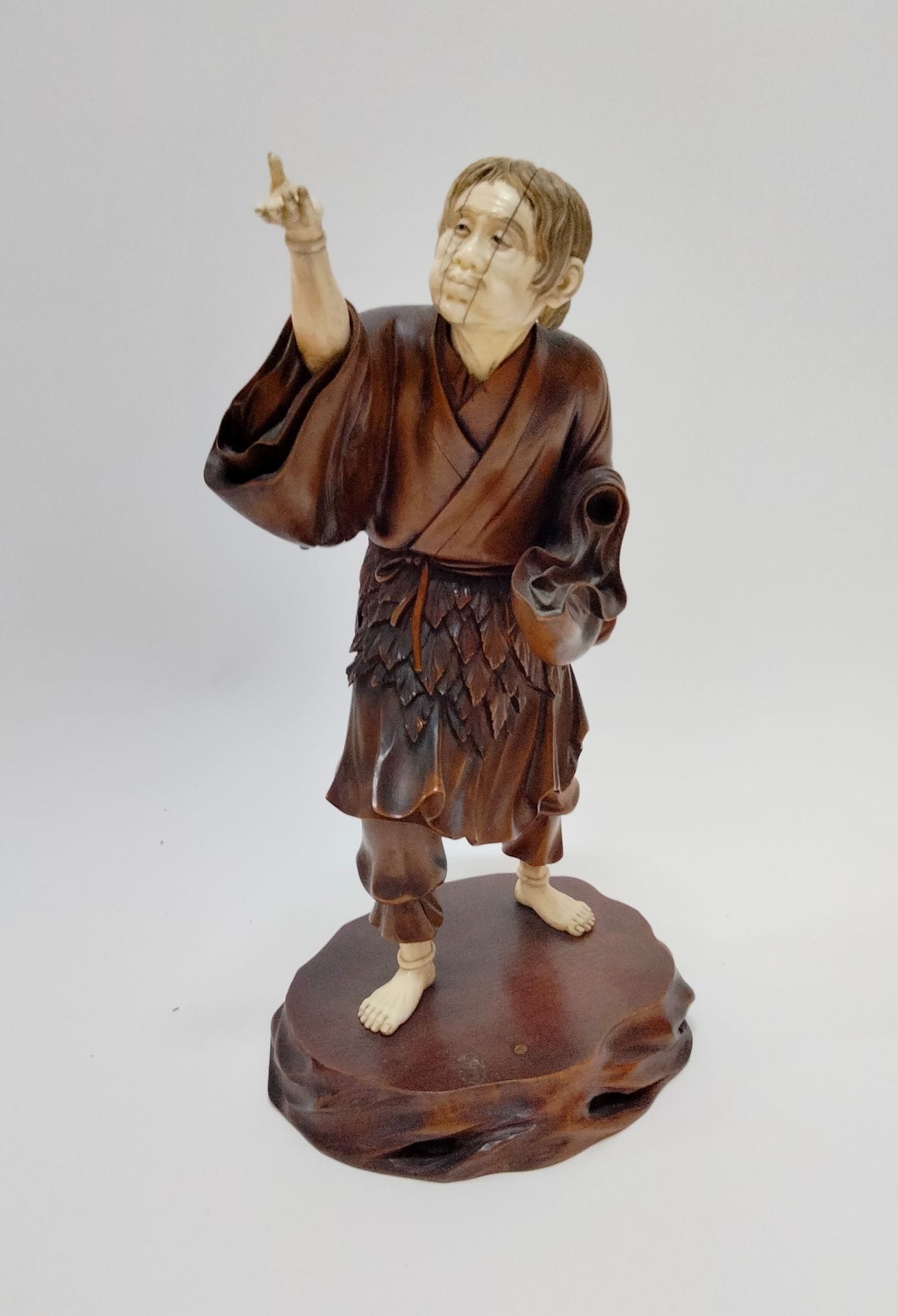 Okimono en bois et ivoire représentant Tekkai sennin (Li tieh kuai en chinois), Japon, époque Meiji (1868-1912)
La tête, l'avant-bras droit, les mains et les pieds ainsi que la gourde sont en ivoire tandis que le reste du corps est en bois. Il regarde sa main ouverte paume dirigée vers le haut de son bras droit levé. Il a les joues gonflées et semble souffler vers sa main. De l'autre main il tenait un bâton noueux disparu. Il est vêtu d'un vêtement usé dont la taille est ceinte d'un entourage de feuilles caractéristique des représentations de sennin et porte à la ceinture une coloquinte servant de gourde. Debout sur une base en bois fixé par deux vis en laiton. Sous cette base un cachet rouge avec la signature Ryôun - 竜雲.
Hauteur : 28 cm ; poids brut : 916 g
Permis CIC n°FR2307503661-K
Gerce au visage, manque une main.