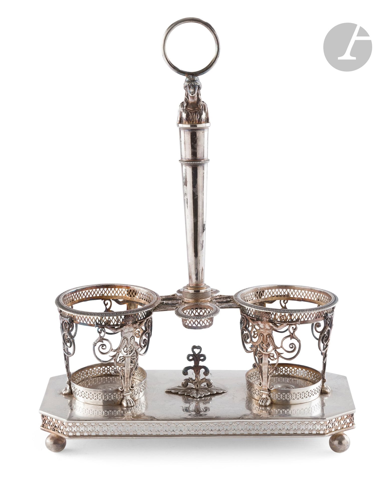 PARIS 1809 - 1818
Monture d'huilier-vinaigrier en argent. Elle repose sur une base rectangulaire à pans et pieds boules. La prise centrale est terminée par un anneau sommant un buste de Janus féminin. Les porte-carafes sont estampés de motifs de putti portant des trompettes. 
Orfèvre : Pierre-Jacques MEURICE 1806 et 1812
Poids : 733 g - longueur : 23,4 cm - largeur : 10,8 cm - hauteur : 31,3 cm