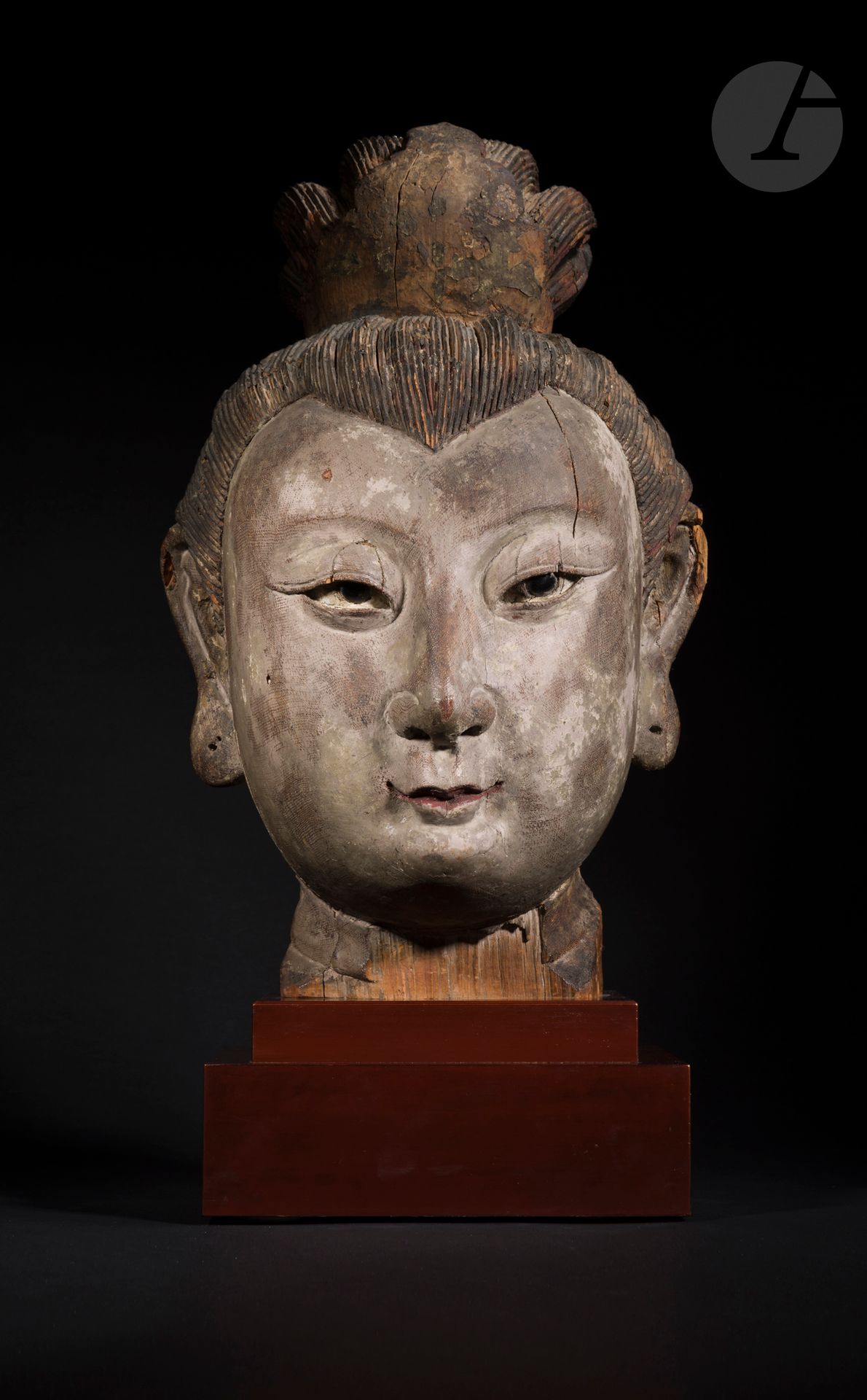 Importante tête en bois de jeune femme noble, Chine, époque Yuan - Ming, XIVe - XVe siècle
Elle porte une coiffure très élaborée tenue à l’avant et à l’arrière par un accessoire serrant le chignon. Les yeux finement sculptés sont incrustés d’une pierre noire figurant l’iris. La bouche est étroite et les lèvres fines sont relevées par une polychromie rouge. Les oreilles ont de larges lobes percés qui ont pu à l’origine être ornés de boucles. Le bois très finement sculpté est recouvert d’une gaze sur laquelle a été appliqué du gesso, très probablement pour solidifier la sculpture réalisée en plusieurs parties.
Hauteur : 46 cm ; h. avec socle : 57 cm
Manques, traces de polychromie.

Provenance : 
Acquise chez Oscar Meyer à Los Angeles entre 1950 et 1971 (copie d’une photographie).