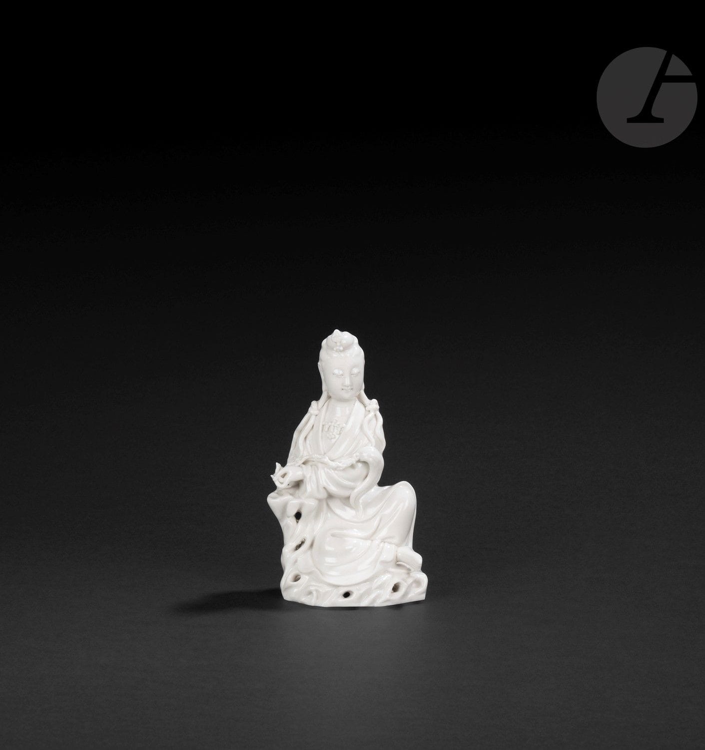 Small Chinese white porcelain guanyin, China, Dehua, 19th - 20th century
She is depicted seated on a pierced rock, wearing a long pleated robe and a pearl necklace. Her serene face features a slight smile, the forehead is adorned with the urna and the earlobes are elongated. The hair is gathered into a small bun, adorned at the front with an ornament, and divided at the back into two long locks passing over the front of the shoulders and knotted to form two new locks. His left arm rests on a rock and his hand holds a fuzi. The naked right foot protrudes from the bottom of the dress. On the back is a molded double-gourd mark, difficult to identify.
Height: 13.5 cm
Foot and top of headdress ornament reattached.