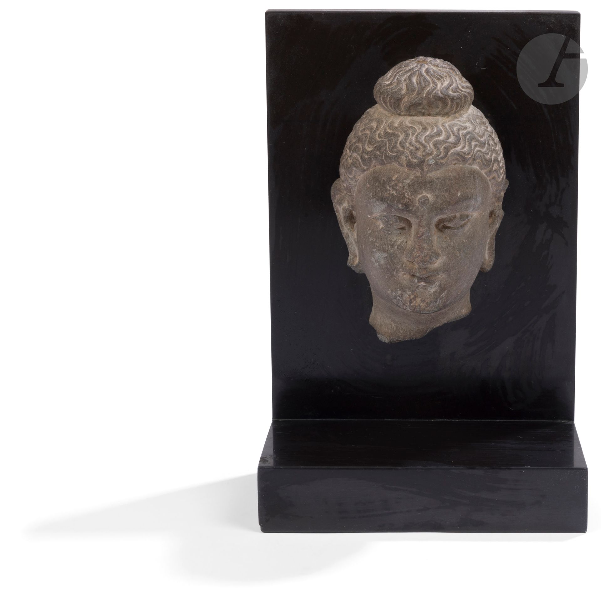 Tête de bouddha en schiste, Art gréco-bouddhique du Gandhara, IIe - IVe siècle
Les yeux légèrement ouverts, la coiffe ondulée ramassée en un chignon bombé ushnisha. 
Hauteur : 14 cm
Montée sur un socle en bois noirci. Côté gauche abimé.