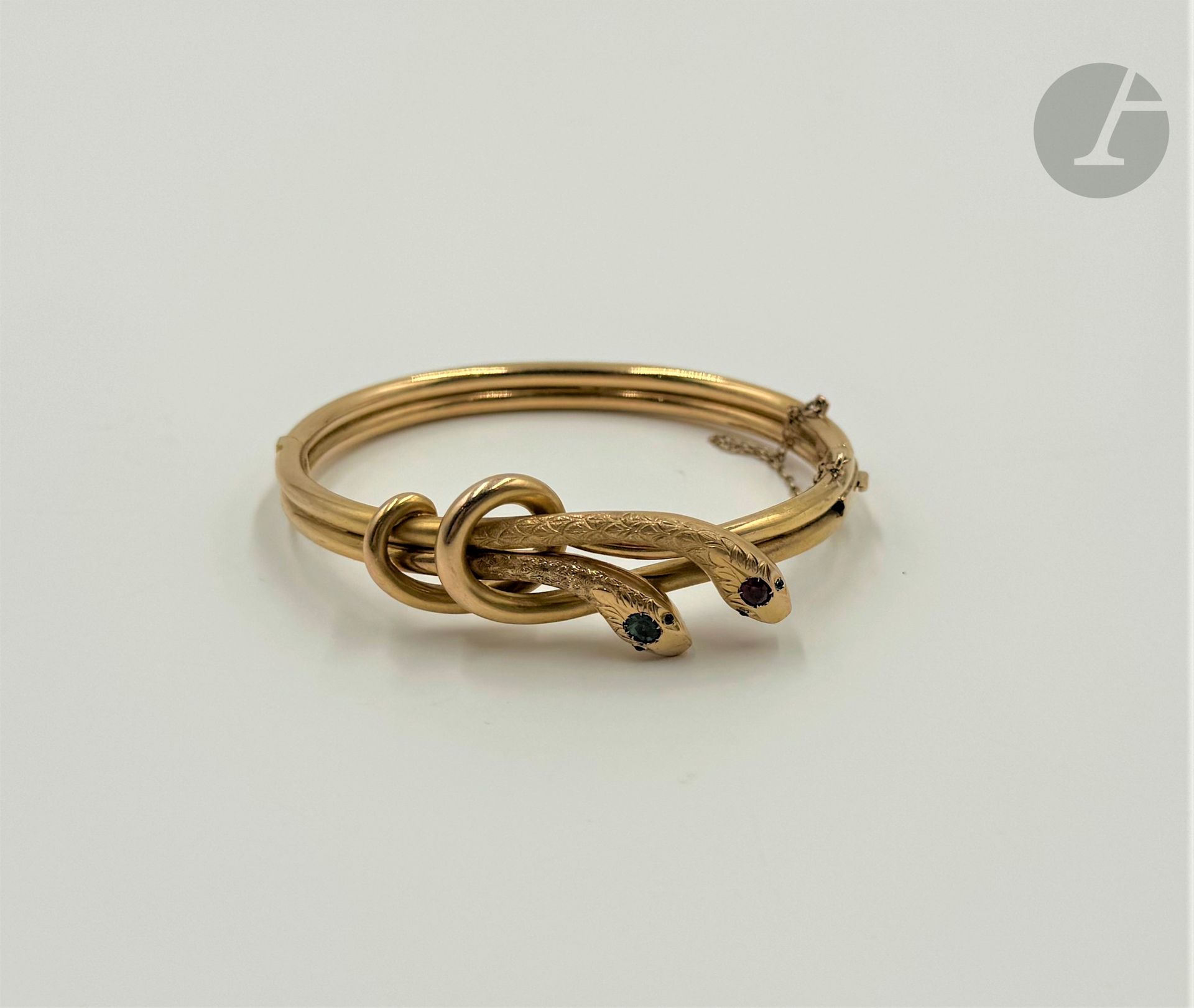 Opening bracelet in knotted 18K (750) gold wire ending in two engraved snake heads, the eyes and top of each head set with synthetic green and red stones. Width: approx. 17 cm. Gross weight: 17.4 g (scratches, chips)