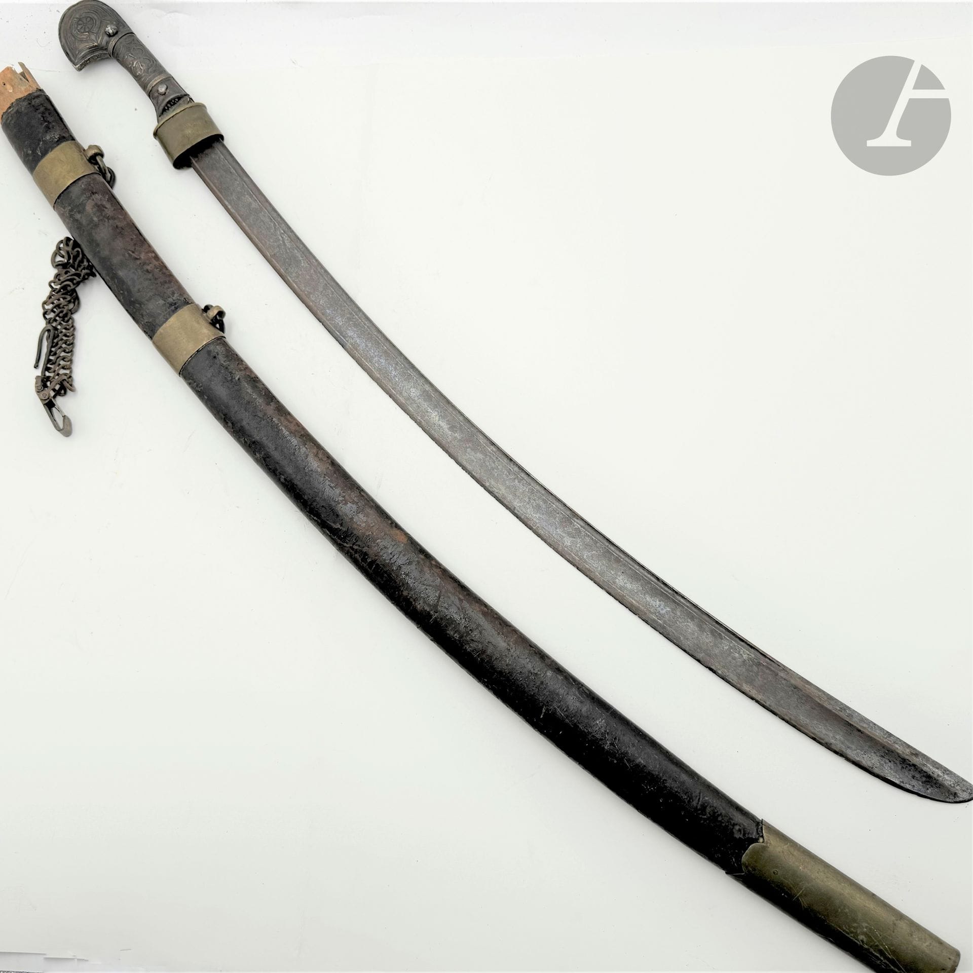 Sabre caucasien «Shashka». 
Poignée en métal argenté niellé à deux rivures. Lame cintrée à dos plat, gouttière et pans creux. 
Avec un fourreau en bois recouvert de cuir à quatre garnitures dont trois avec poinçons en cyrillique. 
Avec une suspente gourmette française. 
E.M. (Usures).