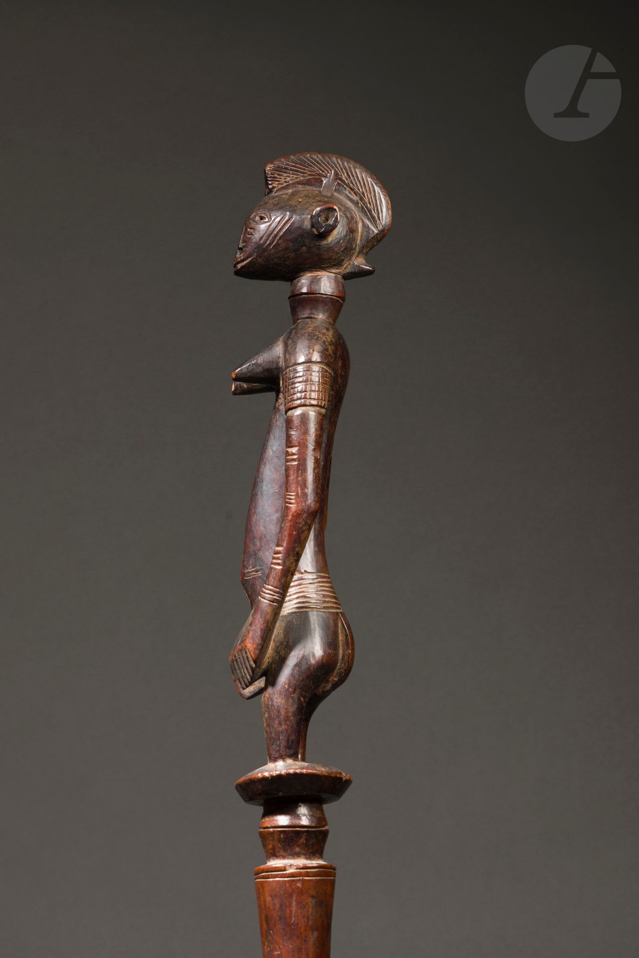 Une canne d’initié de la société du Poro ou un bâton d’escorte, possiblement la canne d’un chef et personnage historique en la personne du Roi Babemba.
D’une plus petite taille que les grandes cannes tefalipitya qui célèbrent le sambali (le champion des cultivateurs) qui sera « récompensé » par une jeune femme non mariée au sommet de sa beauté représentée assise au sommet de ces cannes, la canne senoufo de la collection Jean Roudillon est ornée d’un personnage féminin sculpté en position debout, bien campé, telle une statue déblé.
Il s’agit très certainement d’un bâton d’initié de la société du Poro, ou d’un bâton d’escorte dont l’image féminine évoque les pouvoirs surnaturels des femmes, celle des sandobele, les femmes-devins, qui perçoivent les dangers cachés et passent devant pour écarter les sorts jetés par les sorciers.
Cette canne magnifique à la patine laquée est d’un grand style ancien, dont les grands bras stylisés aux épaules puissamment arquées et les oreilles sculptées en cylindre renvoient sans équivoque aux plus belles statues Déblé de l’atelier dit des maîtres de Sikasso.
Elle a été exposée en 1964 dans trois musées américains lors de l’exposition itinérante Senufo Sculptures from West Africa dont Robert Goldwater, directeur du Museum of Primitive Art de New York, était l’instigateur.
La provenance de cette canne, prêtée par la galerie Le Corneur Roudillon à l’époque, la rattache dans le catalogue de cette exposition au Roi Babemba, personnage historique s’il en est au Mali, ayant succédé en 1893 à son frère Tiéba Traoré, quatrième roi de Kénédougou qui avait mené le royaume à son apogée et fixé sa capitale à Sikasso, où il fit notamment construire son palais pour résister aux attaques de Samory Touré. Le roi Babemba Traoré se suicida en 1898 plutôt que d’être pris, préférant la mort à la honte, après avoir lutté contre l’armée colonisatrice.
C’est forcément Olivier Le Corneur et Jean Roudillon qui ont transmis cette provenance à Robert Goldwater, une provenance qu’ils avaient acquise avec l’objet. Réelle ou non, Goldwater un homme sérieux et historien de l’art devait considérer cette provenance comme authentique pour la valider et la publier, bien qu’aucun autre document ne puisse en attester réellement.
Dans les notes de Jean Roudillon :
« Afrique, Côte d’Ivoire, Senufo
Canne du Roi Babemba de Sikasso
Rapportée par un officier français en1898. 
Publié fig. 135 dans  « The Museum of Primitive Art » par Robert Goldwater, New York, 1964 »
Sénoufo, Côte d’Ivoire
Bois, fer, oxydation d’ancienneté, usures, petit accident à la pointe du sein droit et une restauration indigène en fer au bras droit, très belle et ancienne patine d’usage.
H. : 113 cm
Voir pour les statues-pilon déblé de l’atelier dit des maîtres de Sikasso p. 117 à 137 dans : Senoufo Massa et les statues du Poro, Burkhard Gottschalk, Ed. Verlag U. Gottschalk Düsseldorf 2006
Provenance :
- Ancienne collection Galerie Le Corneur Roudillon
- Collection Jean Roudillon
Expositions et publication :
- Senufo Sculpture from West Africa, Robert Goldwater, Ed. The Museum of Primitive Art, New York, 1964, p. 90 n° 135
- Senufo Sculpture from West Africa, 1963, exposition itinérante à :
- New York, NY The Museum of Primitive Art, du 20 février au 5 mai 1963
- Chicago, IL, Art Institute of Chicago, du 12 juillet au 11 août 1963
- Baltimore, MD Baltimore Museum of Art du 17 septembre au 27 octobre 1963.