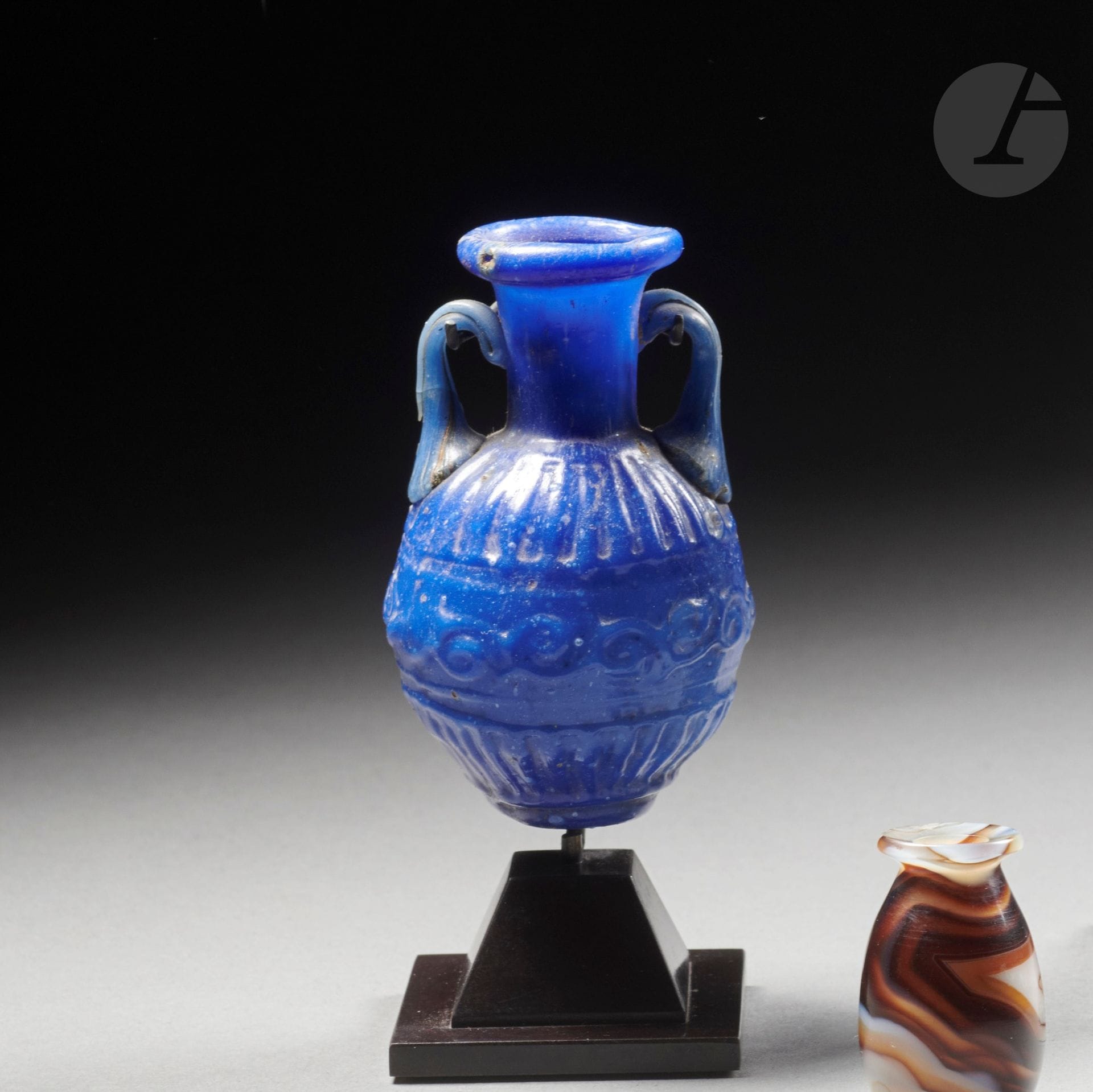 Amphorisque en verre soufflé moulé bleu roi. La panse ovoïde est ornée en son centre d’une frise de volutes, encadrée par un décor de godrons sur l’épaule et en partie basse. Deux petites anses appliquées sur l’épaule et repliées sous l’embouchure. Petit pied cylindrique. 
Verre bleu roi, une anse cassée-collée
Méditerranée orientale, Liban, probablement Sidon.
H. : 7 cm
Note : Sidon est située dans l’actuel Liban et a été un centre de fabrication de matières vitreuses depuis plusieurs millénaires, probablement la ville où est né le verre. À l’époque romaine, Sidon continue d’être active dans l’élaboration de vases en verres. Ces petites récipients soufflé-moulés pouvant avoir diverses couleurs ou décor ont été trouvés dans la région et sont datables du Ier siècle.
Provenance :
Collection Jean Roudillon