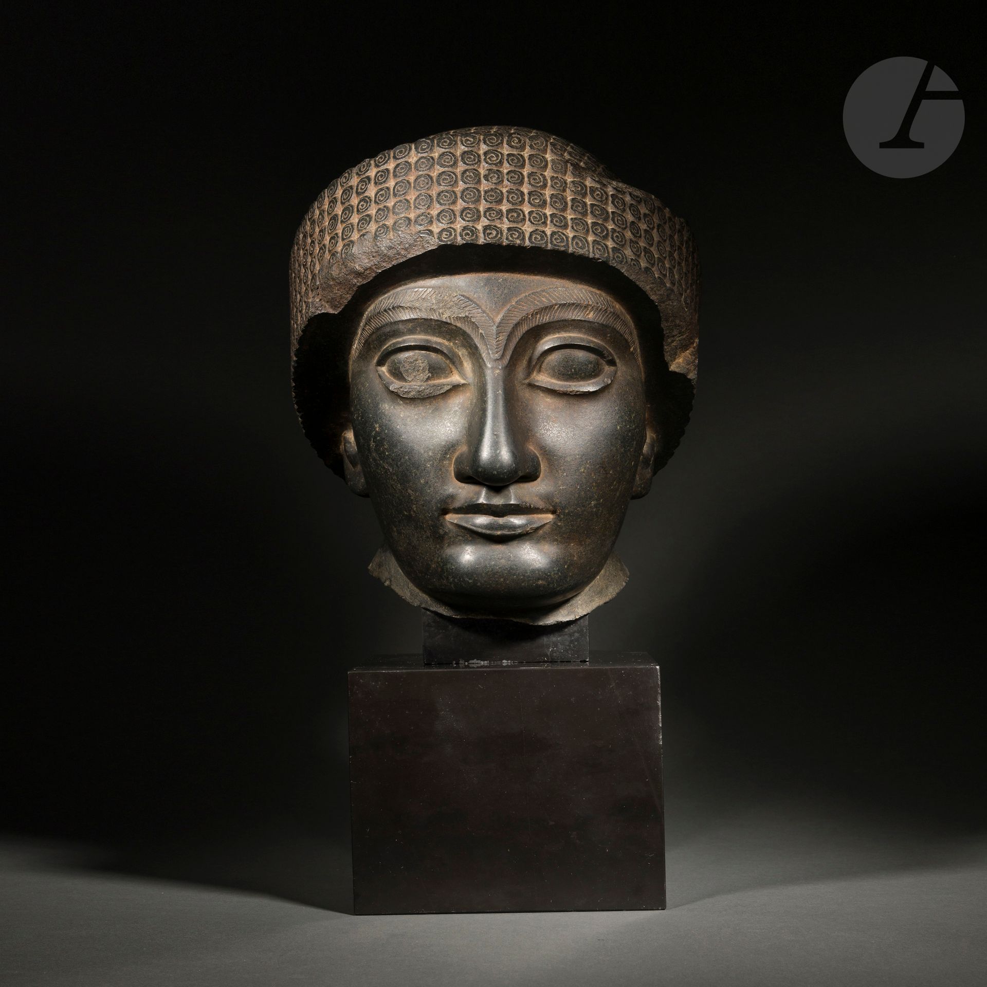 Sculpture en ronde-bosse figurant un portrait de Goudéa, roi de Lagash.
Le souverain est coiffé du bonnet royal à large rebord décoré de boucles. Les paupières ourlées sont surmontées par des sourcils en relief, arqués, striés, se rejoignant sur le haut de l’arête nasale. Les lobes de ses larges oreilles apparaissent sous le bonnet. La bouche s’étire, lui conférant une expression distinctive. Goudéa a maintenu la paix pendant 20 ans sur son royaume. Ces statues témoignent de son règne prospère et de sa piété religieuse. 
Pierre noire polie (diorite), épaufrures et manques visibles, présenté sur un socle en pierre. 
Dans le goût sumérien. 
Note : on recense plusieurs dizaines de statues de Goudéa dans les musées, certaines trouvées dans un contexte archéologique et d’autres acquises sur le marché de l’art.
H. : 33 cm
Provenance :
- Collection Ernest Ascher
- Collection Jean Roudillon