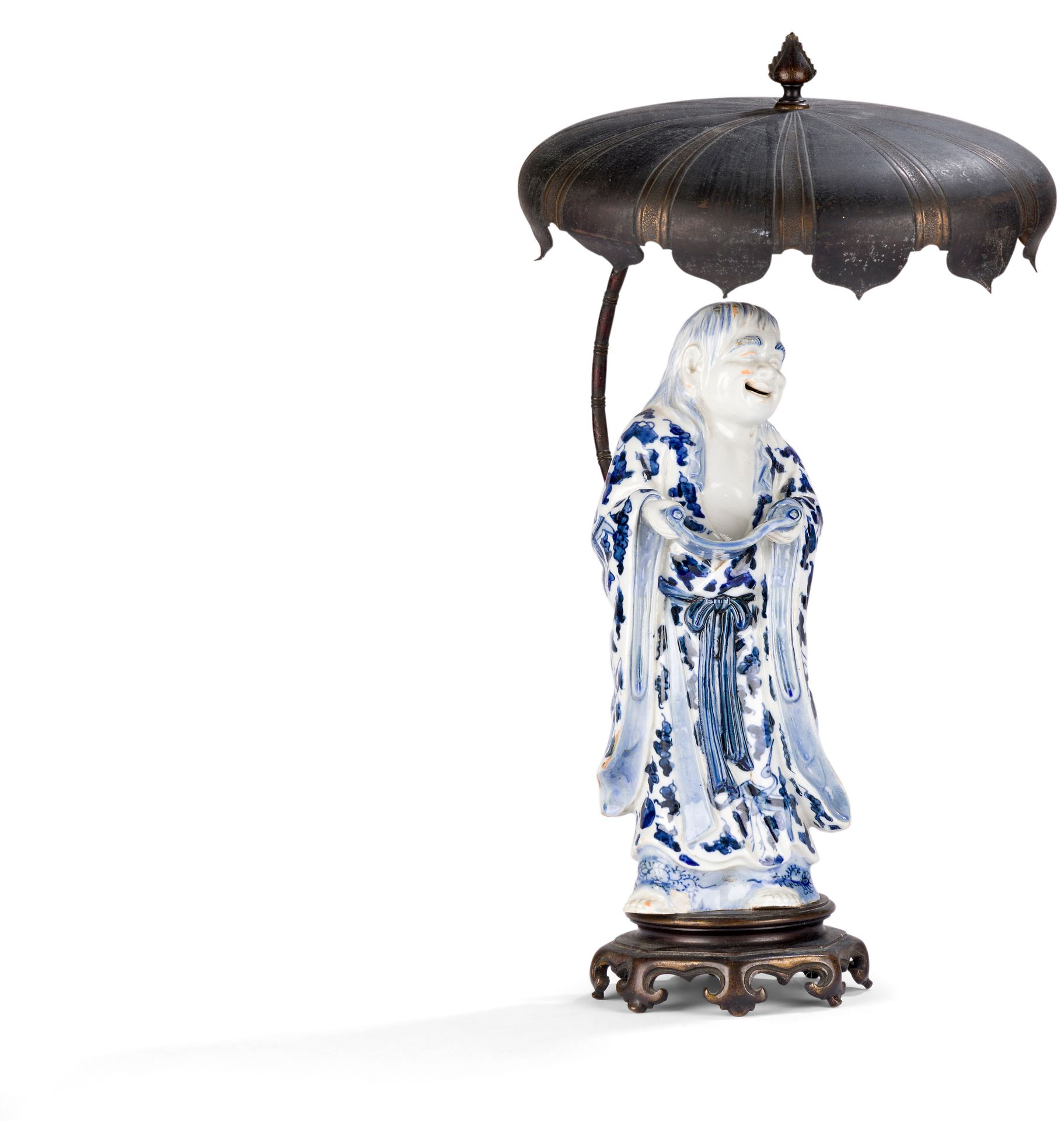 Personnage représentant Kanzan, en porcelaine bleu et blanc, monté en lampe, Japon, XIXe siècle
Kanzan, vêtu d'un kimono à décor émaillé en bleu sous couverte à décor de tsuru (grues à tête rouge), nuages stylisés et objets précieux, tient dans ses mains un rouleau calligraphié qu'il regarde avec un sourire. Il est monté sur une base octogonale à huit pieds en alliage de cuivre, à l'arrière de laquelle monte une tige courbe imitant un bambou finissant en large parasol.
Hauteur : 28,2 cm
Petit éclat.

Kanzan, dont le nom signifie "montagne froide", est un poète zen qui cherche son inspiration dans la nature. Bien qu'ils se ressemblent énormément, on le distingue facilement de son alter ego Jittoku, car il tient toujours dans ses mains un rouleau de parchemin.