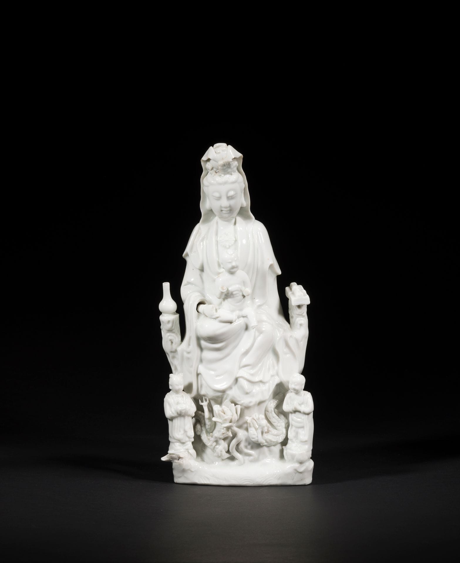 Large Chinese white porcelain Guanyin, China, Dehua, 18th century
Seated, holding a child, with her right leg bent and her left leg dangling in a position known as vamardhaparya?ka. On one side of her throne, a bottle of lustral water on her right, a scroll and sutra books on her left. At his feet, his two acolytes LongNü, holding the Pearl of Light, and ShanCai TonZi, hands in adoration. Between them, on a terrace, a lotus flower surrounded by two facing dragons.
Height: 36.3 cm
Minor accidents.