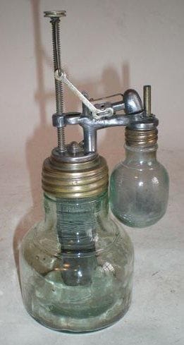 Allumoir d'office automatique, à pile chimique. Modèle tout en verre, comprenant une pile chimique type Grenet, à charbons-zinc, surmontée d'un dispositif manuel à poussoir, permettant l'allumage (par l'incandescence d'un fin fil de platine) d'une petite lampe à pétrole ou à alcool, juxtaposée au corps de la pile. Ensemble curieux, complet et en bon état, c. 1900