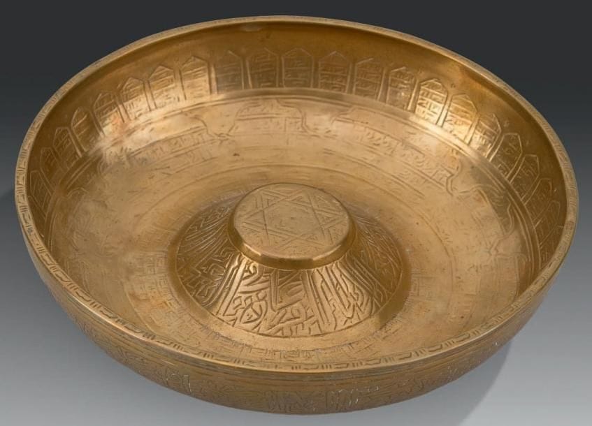 Coupelle prophylactique, Jezireh, XVIe-XVIIIe siècle Coupelle à ombilic en laiton, gravée à l'intérieur de cartouches entrelacés et arcatures meublés de pseudo-inscriptions autour d'une étoile à six branches, et à l'extérieur d'un large bandeau pseudo-épigraphié autour d'une rosette centrale. D.: 18,5 cm (Forte érosion des inscriptions) Provenance: ancienne collection Toulouse-Lautrec, vente publique, Paris, Me F. de Ricqlès, exp. Cabinet Soustiel- David, Paris, 25 septembre 1998, lot 8.; collection J.M.G., Paris L'usage de ces coupelles prophylactiques était très répandu dans le monde musulman, jusqu'à une période relativement récente. Il est parfois difficile de leur attribuer une datation précise, tant l'usage en a érodé les motifs. Pour d'autres coupelles du même type voir le catalogue de l'exposition A l'ombre d'Avicenne. La médecine au temps des califes, Paris, IMA, 18 nov. 1996 au 2 mars 1997, p. 226-227