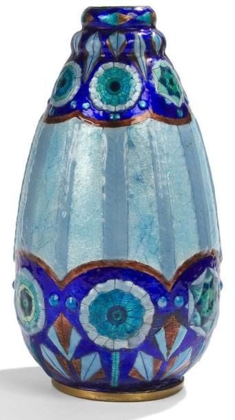 Jules SARLANDIE (1874-1936) à Limoges
Très rare vase obus reposant sur talon et à col en gradins. Épreuve en laiton au décor tournant, de bandes verticales et de fleurs très stylisées dans des réserves vers la base et vers le col, réalisé aux émaux translucides et opalescents sur fond d?un paillon argent, en partie, coloré bleu et turquoise. Défauts ou restaurations.. Circa 1925. Signé J. SARLANDIE (au J&S entrelacé) et situé Limoges vers la base. Hauteur: 28 cm