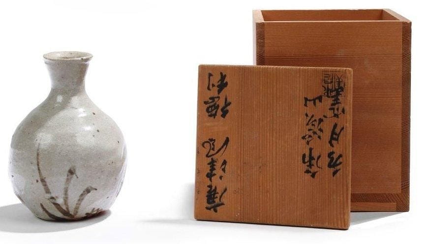 Arakawa TOYOZO (1894-1985)
Potier japonais élevé au rang de Trésor National vivant en 1955. Bouteille à saké, de style Karatsu, en grès émaillé au discret décor brun kaki vers la base. Vendue dans son coffret en bois sur lequel figurent les inscriptions en idéogrammes suivantes: Karatsu-fuu (style Karatsu) - Tokuli (carafe pour saké) - Kokeï-zan (montagne Kokeï) - Suiguétsu-Gama (la désignation du récipient).. Porte une étiquette papier numérotée 44 sous la bouteille. Hauteur bouteille: 13 cm. Dimensions coffret: 15,5 x 12 x 12 cm