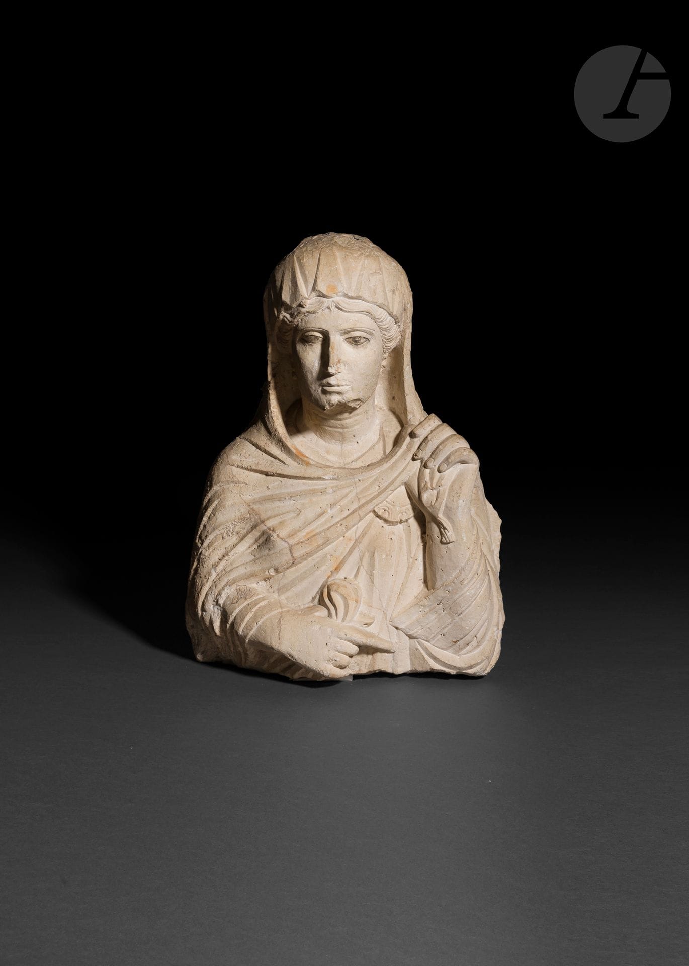Buste féminin sculpté en haut-relief 
La défunte porte un voile sur sa tête qui recouvre une coiffure méchée frontale, séparée par une raie médiane. Ses yeux en amande sont soulignés par de la polychromie noire mais ne sont pas incisés. Son bras gauche est relevé, comme accoudé, tandis que sa main est tendue pour retenir son voile, son bras droit, rabattu contre sa taille tient également un morceau d’étoffe. Elle porte des boucles d’oreilles constituées de trois perles globulaires. Elle est parée d’une broche circulaire ornant sa poitrine, accrochée à sa tunique. 
Calcaire, polychromie noire pour les yeux. Éclats visibles. Trois veines parcourent le buste. 
Syrie, Palmyre, IIe siècle.
Hauteur : 53 cm 

Provenance : 
Ancienne collection de Monsieur Jérôme Carcopino (1881-1970), archéologue, épigraphiste et historien des antiquités latines. En poste à Alger à partir de 1913 où il est nommé directeur du Musée des Antiquités algériennes et Inspecteur des Antiquités. Puis par descendance.