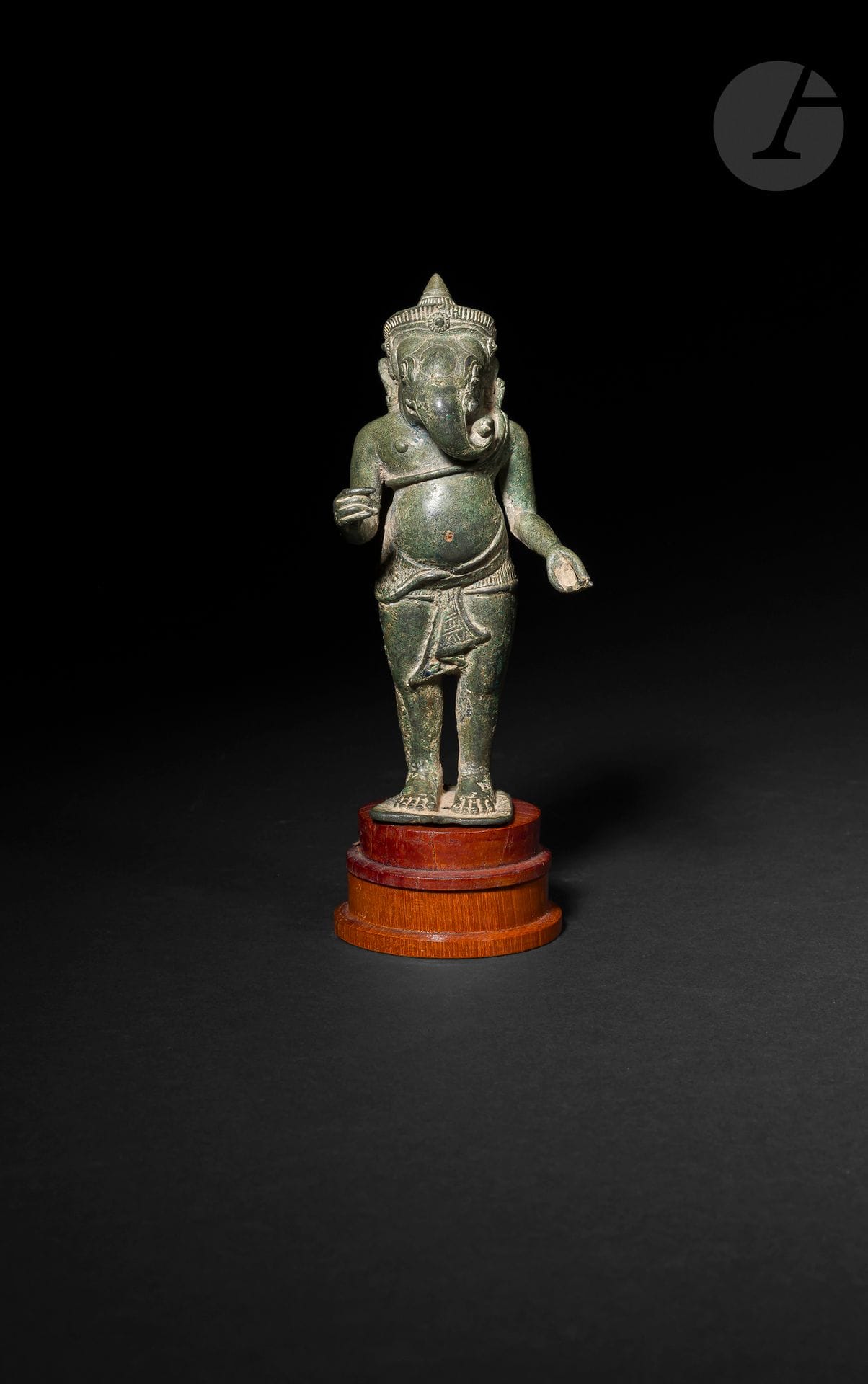 Ganesh debout en alliage de cuivre à patine verte, art khmer, XIe - XIIIe siècle
Représentée debout en samapada, la divinité à tête d’éléphant est vêtue d’un court sampot noué autour de la taille en queue de poisson. Elle tient une défense cassée dans la main droite et probablement un gâteau dans la gauche. Un serpent est enroulé autour de son torse dont la tête est positionnée près de l’oreille gauche. Les petites oreilles parées de bijoux, le front bombé, les yeux aux paupières soulignées, les plis de la bouche et la trompe proéminente enroulée et tournée vers la gauche sous laquelle apparaît une petite défense sont très finement rendus contribuant à procurer une puissante expression à la divinité. La tête du Ganesh est couronnée et surmonté d’un chignon conique. Base munie d’un téton conique en alliage de cuivre. 
Hauteur : 15 cm ; poids : 381 g
Socle en bois.

Provenance : 
- Ancienne collection Evrard de Rouvre (1923-1979). Par descendance. Avec la fiche manuscrite du collectionneur.
- Selon les notes du collectionneur, acquis en décembre 1973 (Ming Siam ?).