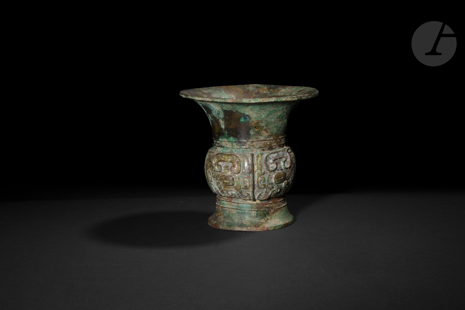 Vase à vin rituel zun en alliage de cuivre, Chine, Époque Zhou, XIe siècle avant J.-C.
De forme circulaire à col et pied évasés. La panse médiane légèrement renflée est ornée d’une frise moulée figurant deux masques de taotie, créature mythique stylisée et symétrique formée par des yeux globuleux, une crête nasale en relief, des cornes involutées et des crocs sans mâchoire. Deux bandes étroites sans décor encadrent la panse. Une inscription à trois caractères moulée dans le fond intérieur du vase : 乍且甫. Belle patine de fouille, oxydation verte.
Hauteur : 18 cm ; diamètre : 19 cm ; poids : 1762 g
Réparations.

Provenance : 
Ancienne collection Nourhan Manoukian (1906-1993). Par descendance.