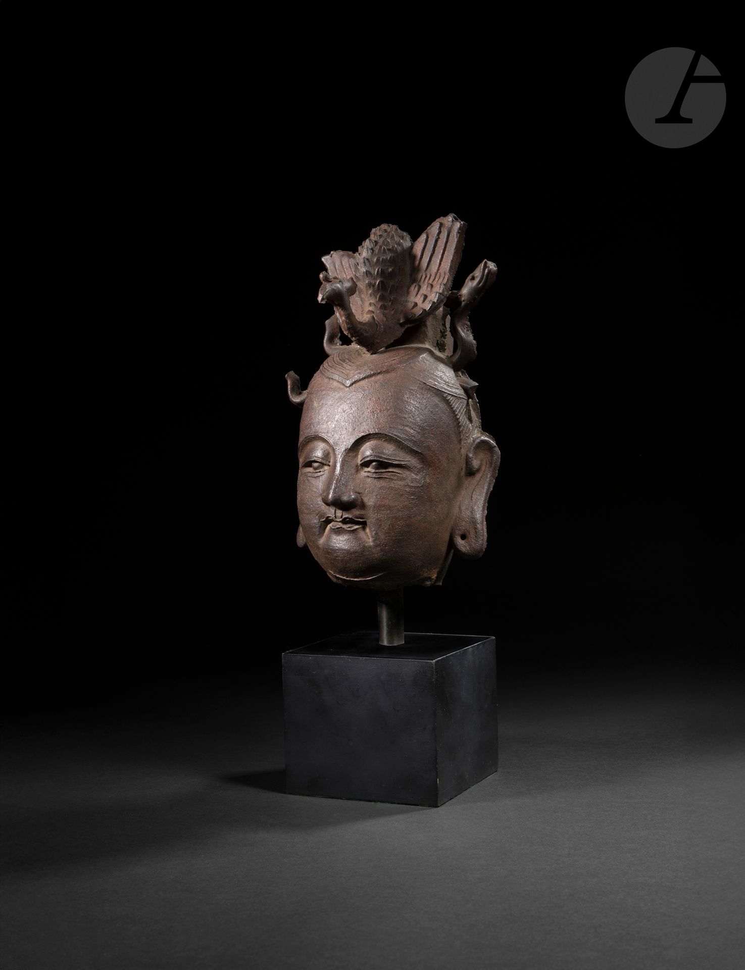 Cast-iron Guanyin head, China, Yuan - early Ming period
Finely molded face, open eyes with eyelids underlined by arched eyebrows, straight nose above a small mouth with hemmed lips. Well-combed hair peaks at the top of the forehead, covers the temples and the back of the head and ends at the nape of the neck. The earlobes are drooping and pierced. The Guanyin's head is surmounted by a fenghuang with outstretched wings and finely rendered plumage. The bird was originally surrounded by long dragons undulating in pairs on either side, but only one on the left is intact.
Height: 21 cm
Traces of mold on the back and sides of the head. Missing parts, notably the tail of the fenghuang and an accident above the left ear. Base.

Provenance: 
- Former Evrard de Rouvre collection (1923-1979). By descent. With the collector's handwritten record. 
- According to the owner's notes, acquired from Galerie Moreau-Gobard, Paris.