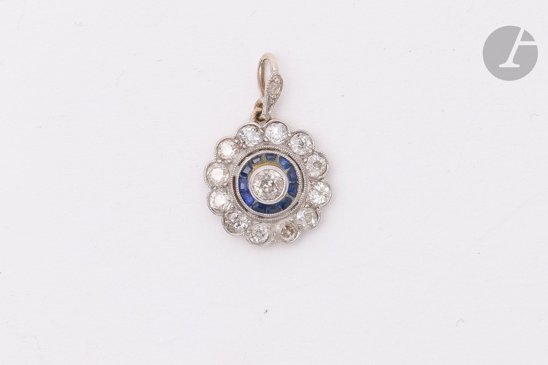 Pendentif fleur en or 18K (750) et platine, le coeur serti d'un diamant rond de taille ancienne dans un double entourage de saphirs calibrés et de diamants ronds de taille ancienne plus petits. Diamètre : 1,4 cm environ. Poids brut : 1,9 g (chocs, égrisures)