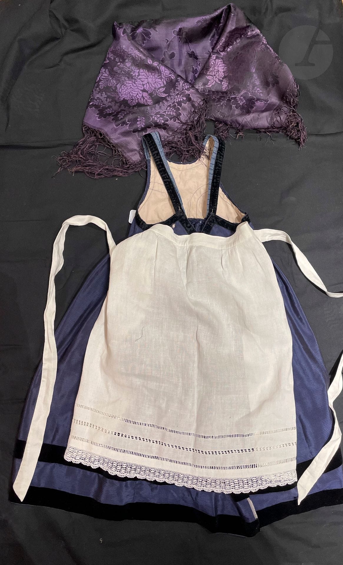 Robe en soie bleue, tablier à cordelière de Berstett, et fichu, Alsace, fin XIXe siècle. Robe entière à bretelle, soie bleu myosotis et galon de velours noir ; tablier de fête en linon blanc à jours et un foulard en soie damassée teint en pièce en prune-parme à franges.