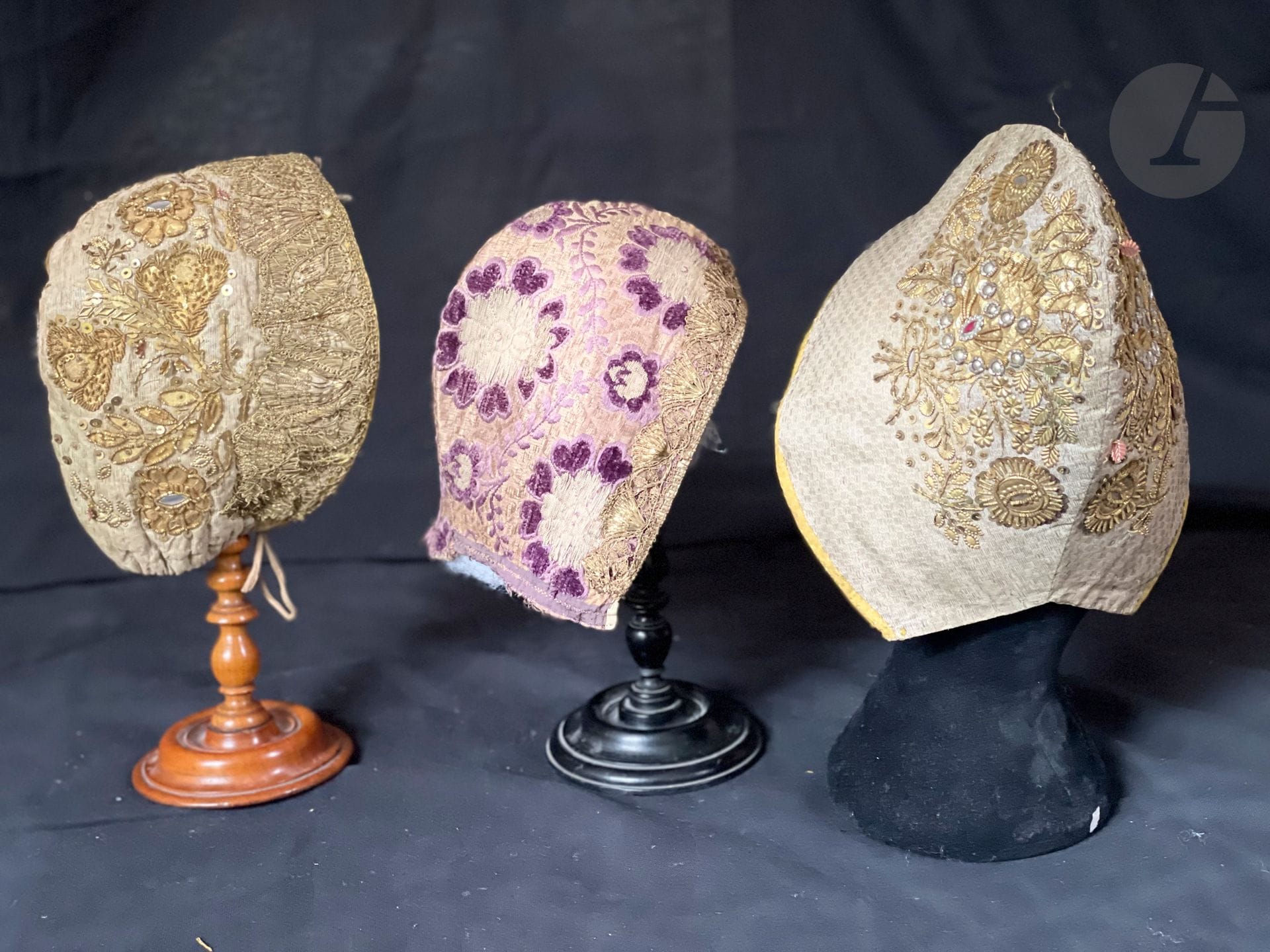 Réunion de 3 coiffes d'apparat, fin XVIIIe- 1er tiers du XIXe siècle. Deux en lampas ou drap d'argent richement brodé de bouquets en cannetille, paillons, verroterie et clinquant, dentelle d'or sur l'une ; la 3e en en lampas soie et velours à fleurons lie de vin, dentelle de lame d'or sur la passe.
